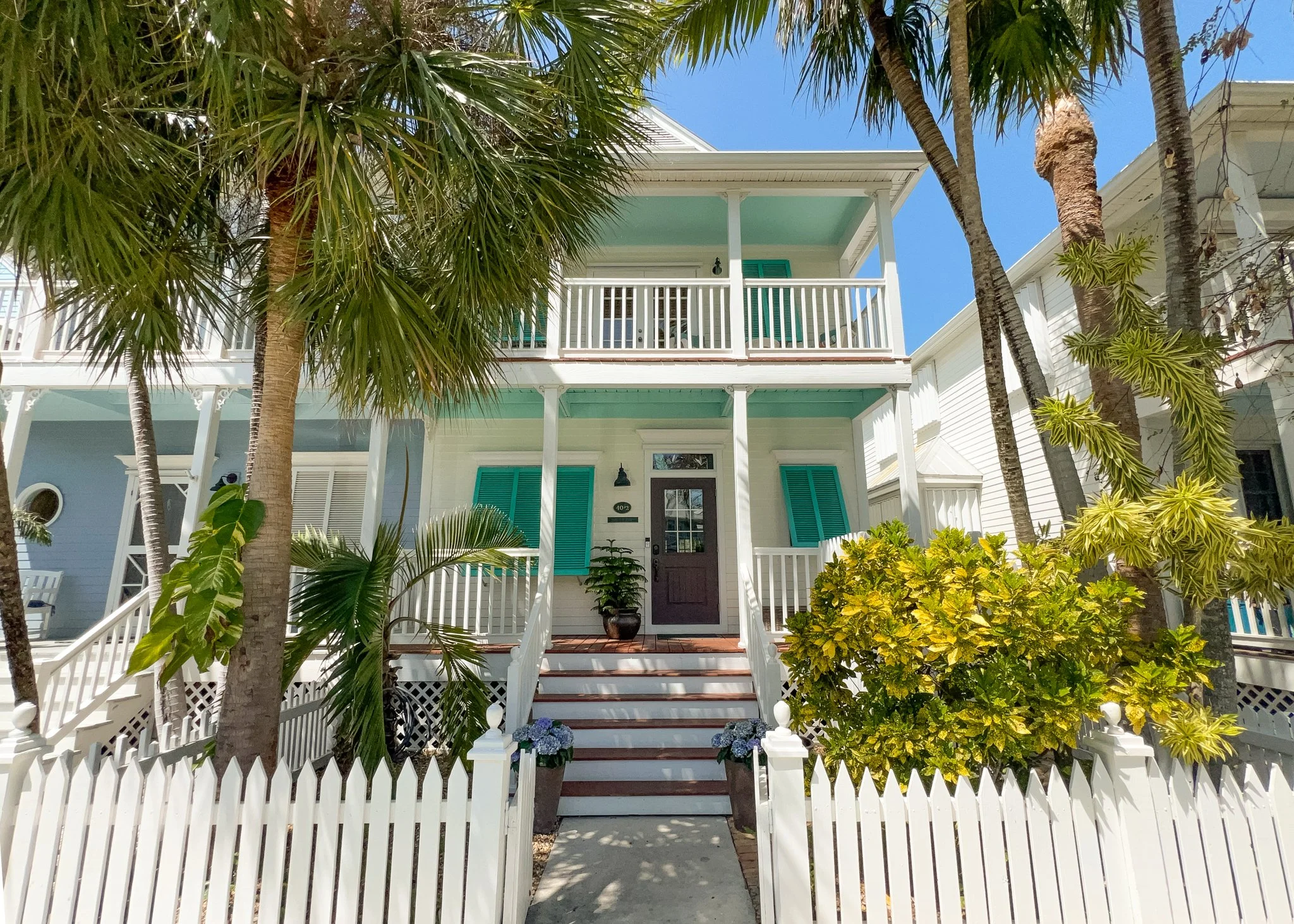 A white, two-story house with turquoise shutters, a front porch, and a white picket fence, surrounded by palm trees and tropical plants under a blue sky.