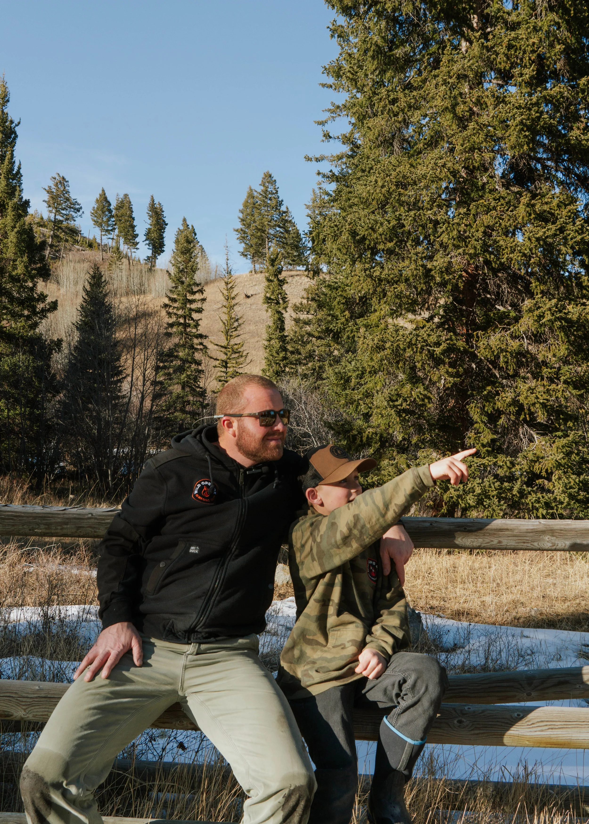 A man and a boy sitting on a wooden fence in a forested area, with the boy pointing into the distance, surrounded by evergreen trees, under a clear blue sky.