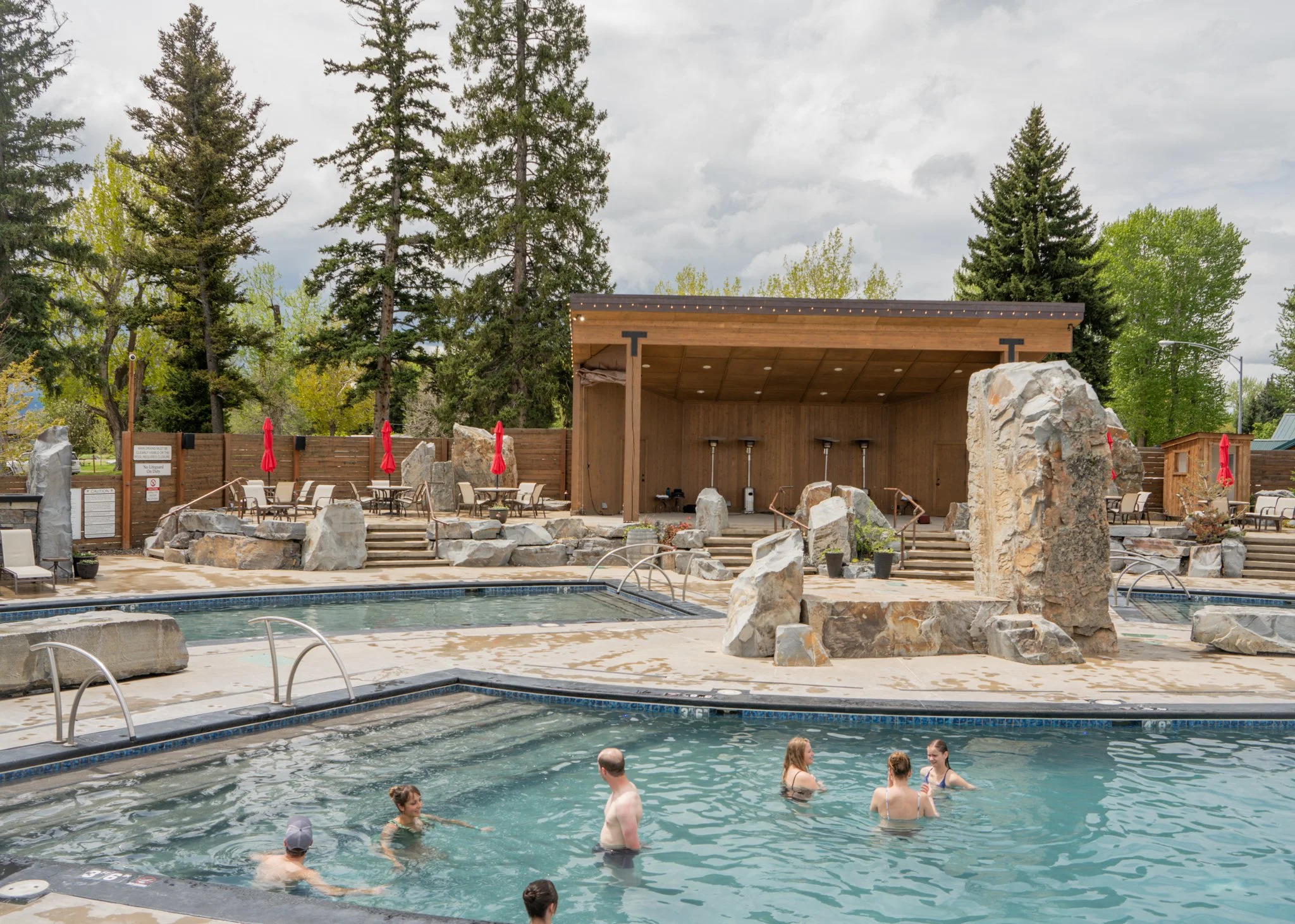 Public outdoor swimming pool area with a group of people swimming and relaxing in the water, surrounded by rock formations, trees, lounge chairs, and a wooden stage or shelter with red umbrellas in the background.