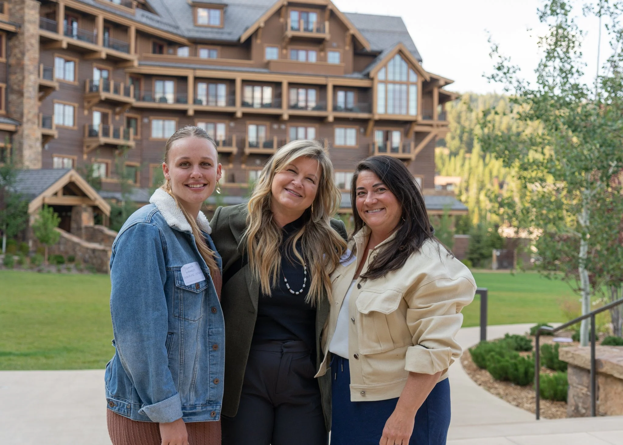 Three women smiling outdoors in front of a large wooden building, with green trees and grass in the background.