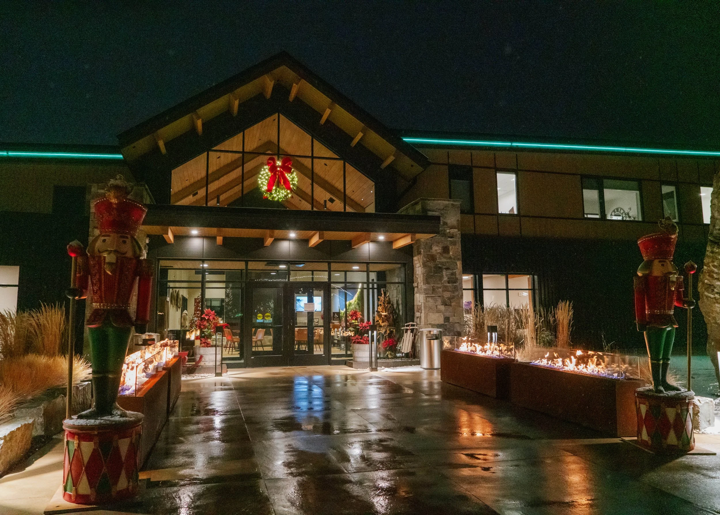 A nighttime view of a modern building decorated for Christmas, featuring a large wreath with a red bow inside the glass entrance, two nutcracker statues on either side, and fire-filled planters along the walkway.