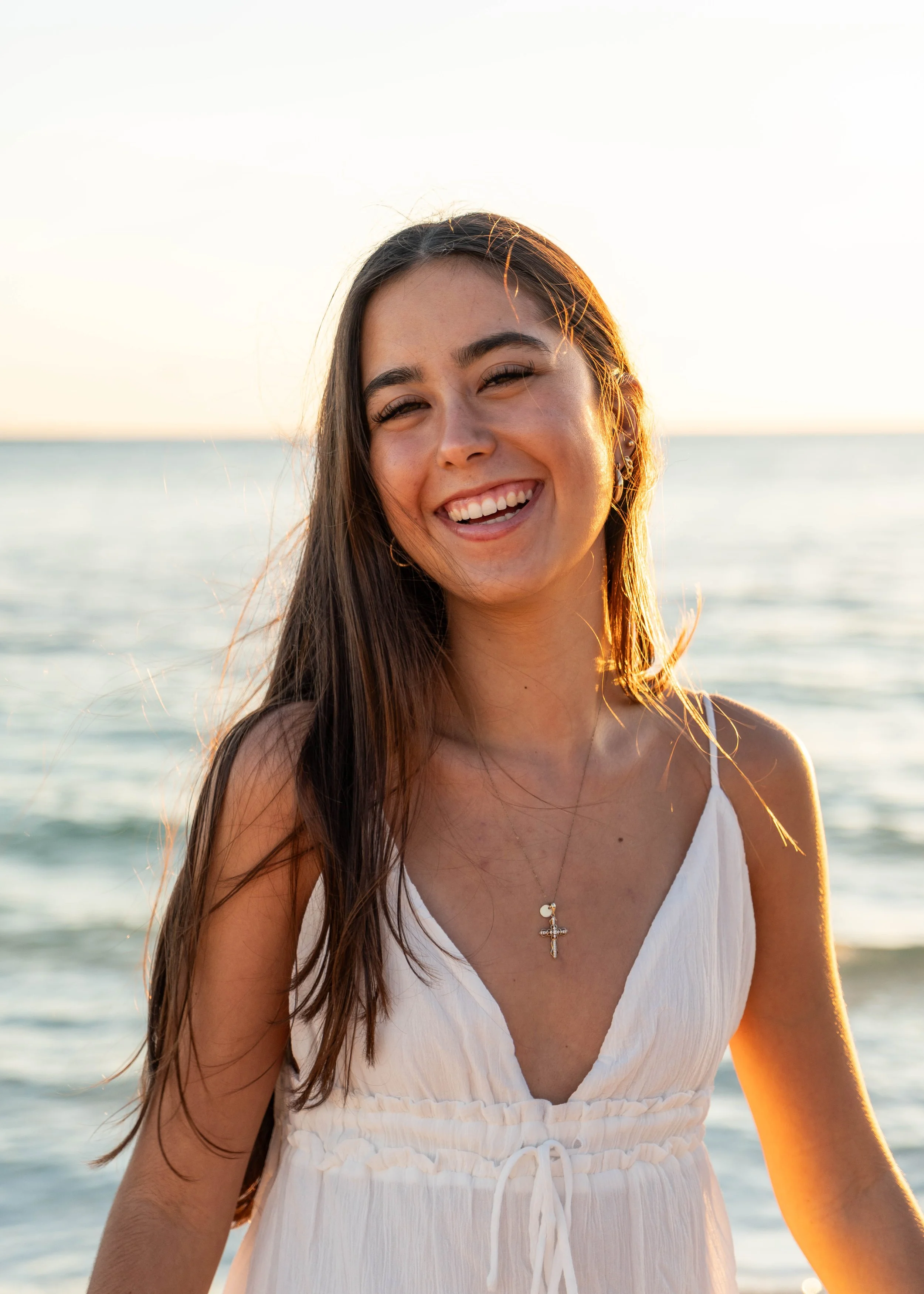Young woman smiling at the camera on the beach during sunset, wearing a white dress and a cross necklace.