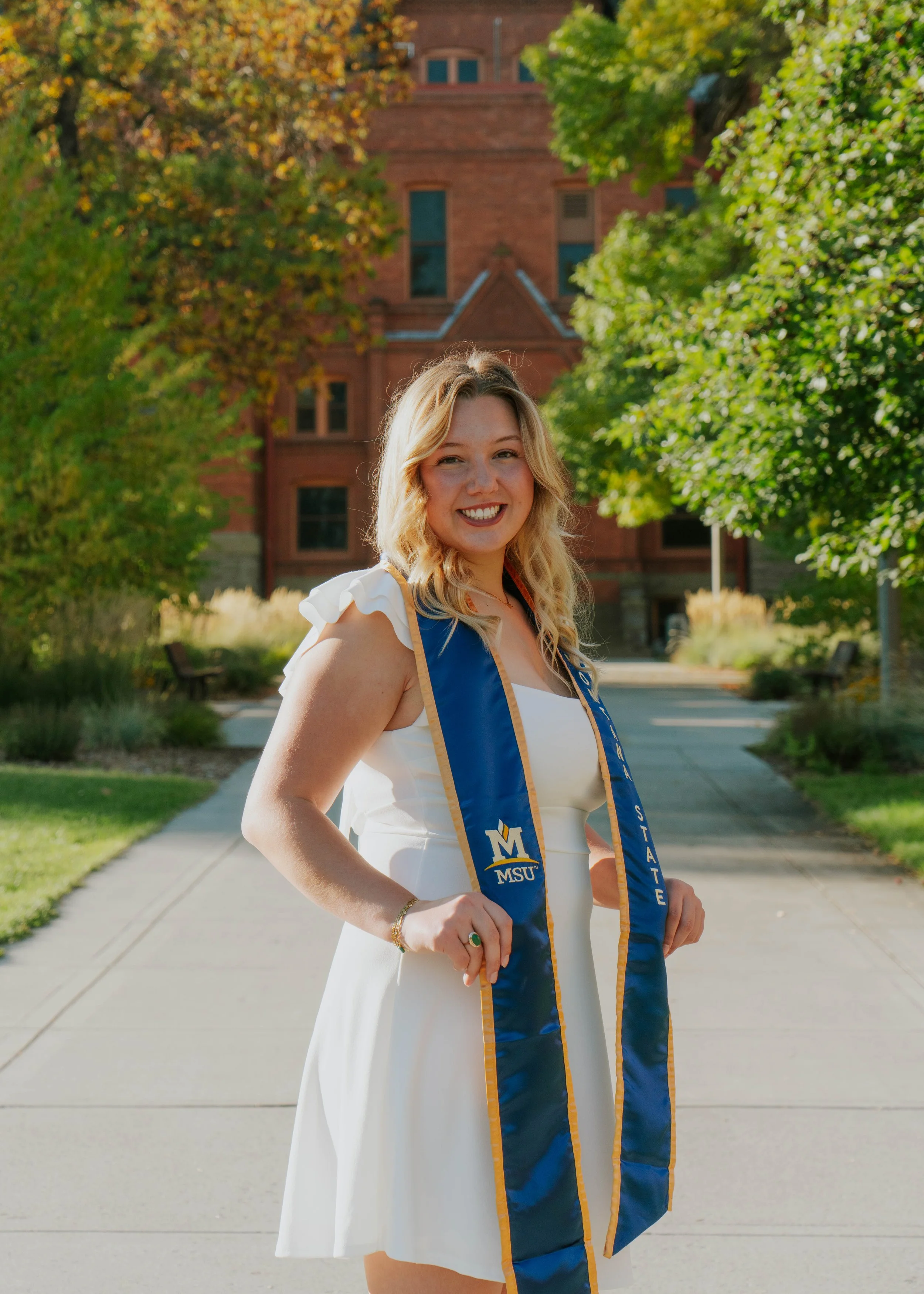A young woman with blonde hair smiling outdoors on a college campus, wearing a white dress and a blue and gold sash that reads 'MSU' and 'State', with green trees and a red brick building in the background.