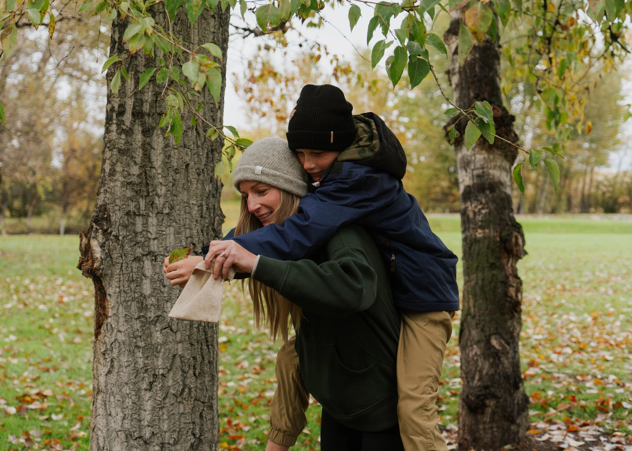 A woman and a young boy in outdoor clothing and knit hats playing near trees in a park during fall, with the boy giving the woman a small bag and the woman smiling.