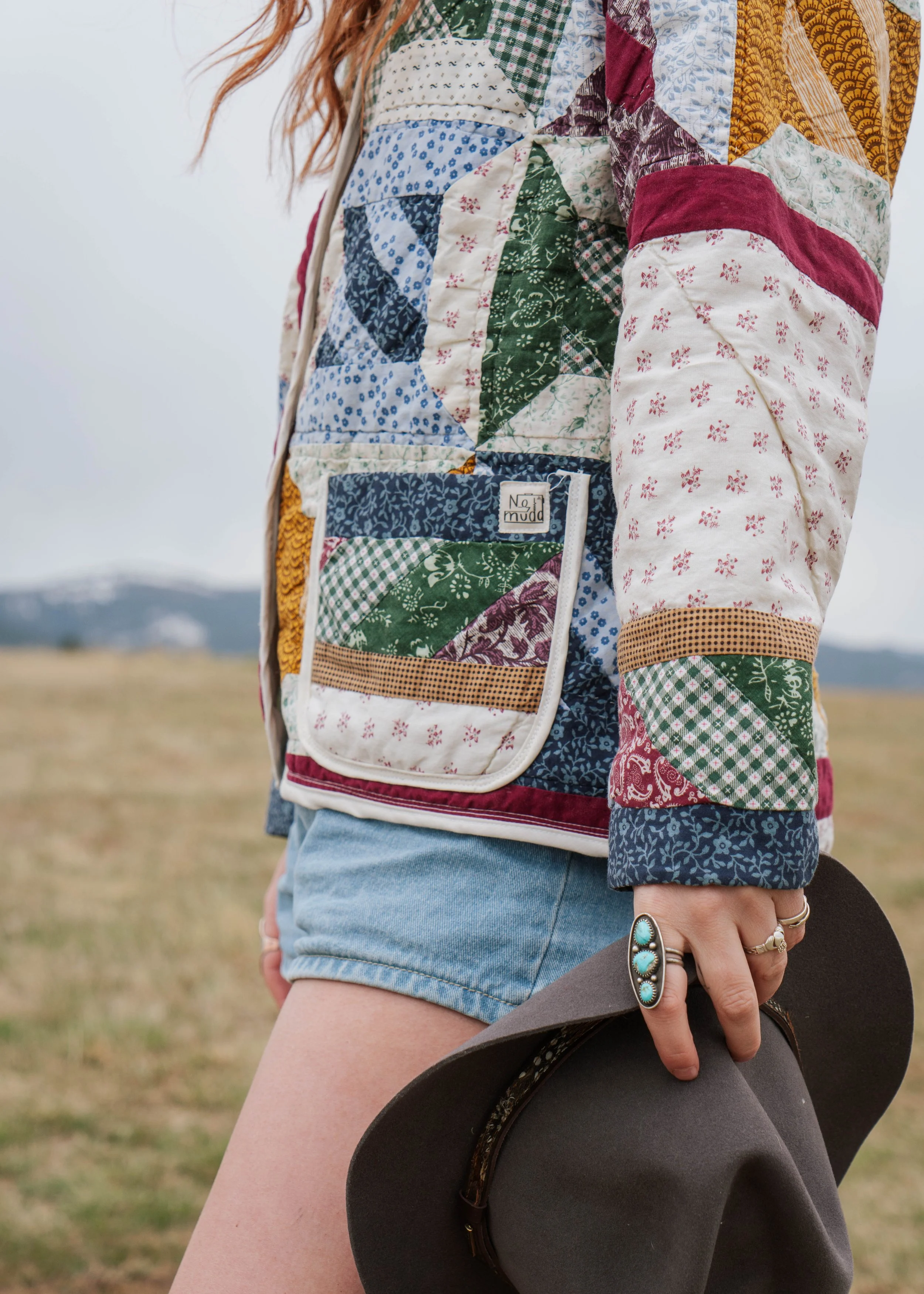 Close-up of a woman wearing a colorful patchwork quilted jacket, holding a gray hat, with a grassy field and mountains in the background.