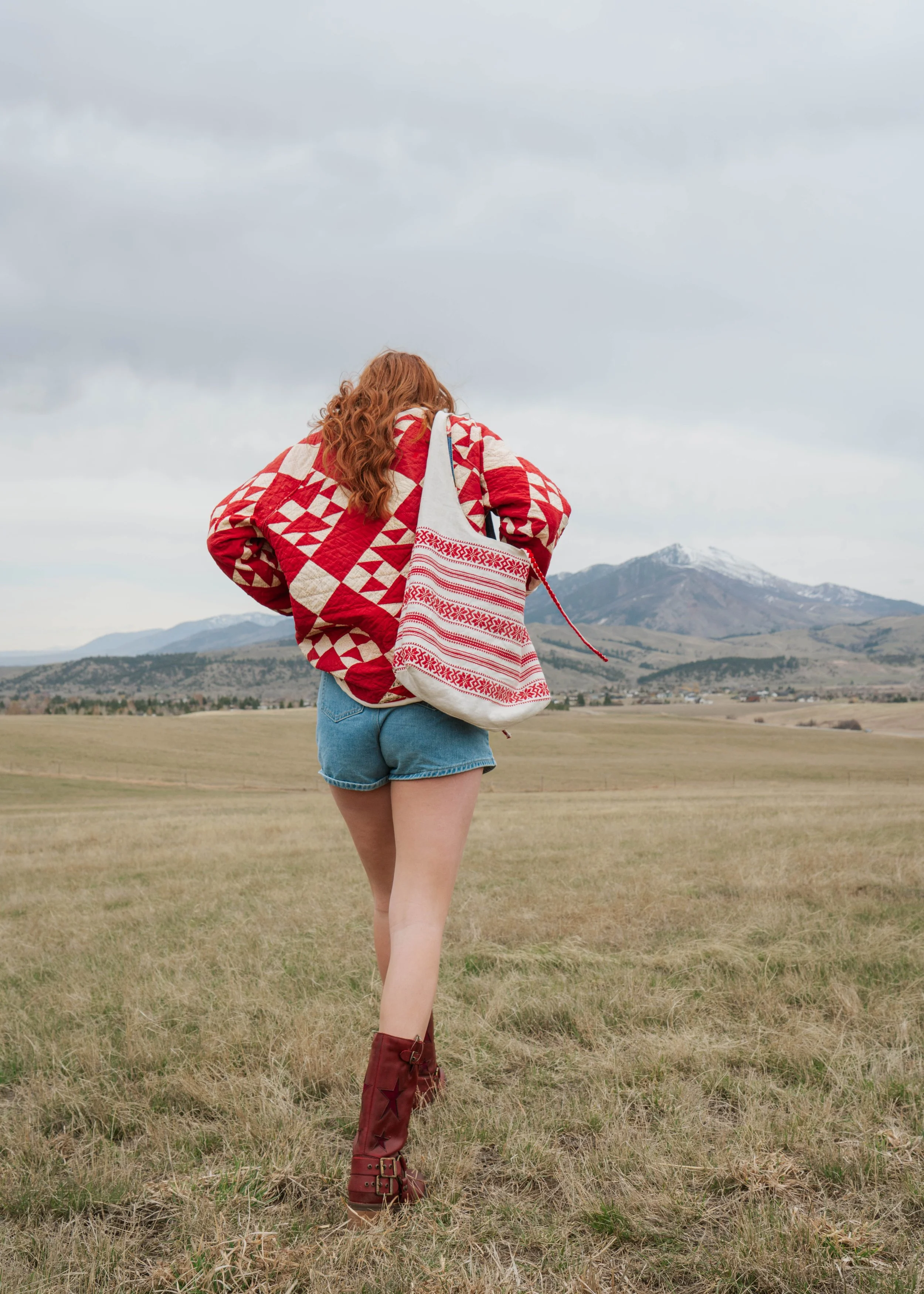 A woman with red, curly hair walking through a grassy field with mountains in the background. She is wearing a red and beige patterned quilted jacket, denim shorts, and red boots, carrying a striped red and white tote bag.