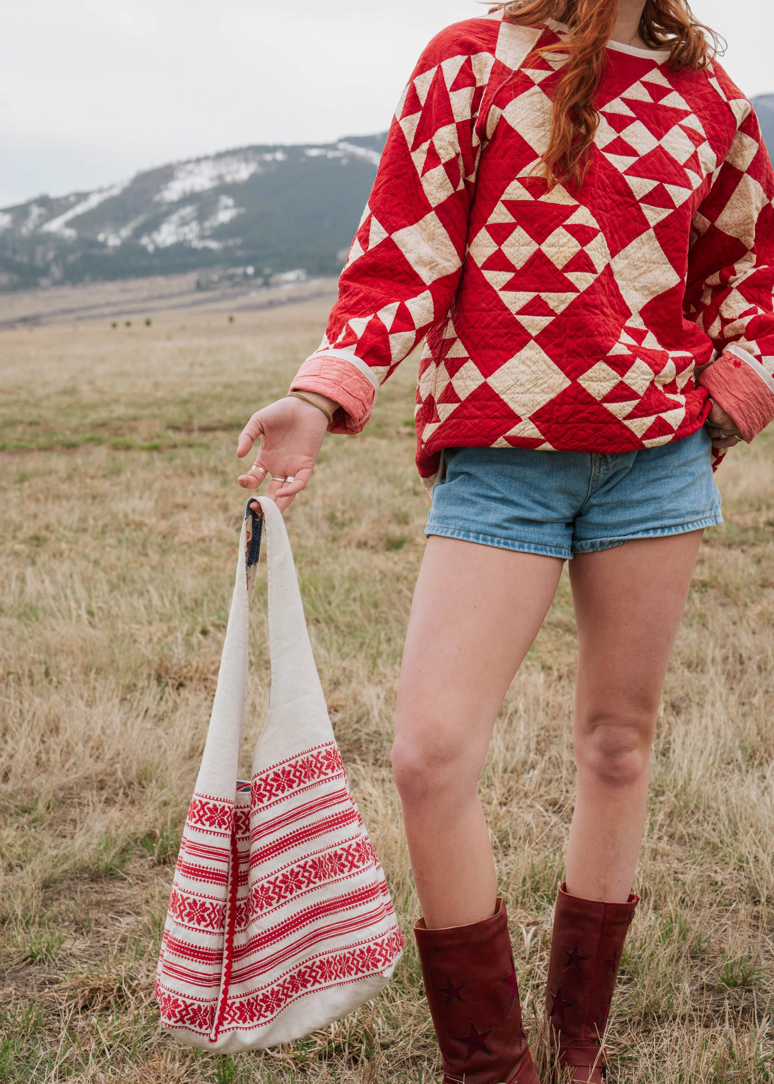 A woman wearing a red and beige quilted jacket and denim shorts holding a white bag with red embroidery in an open field with mountains in the background.
