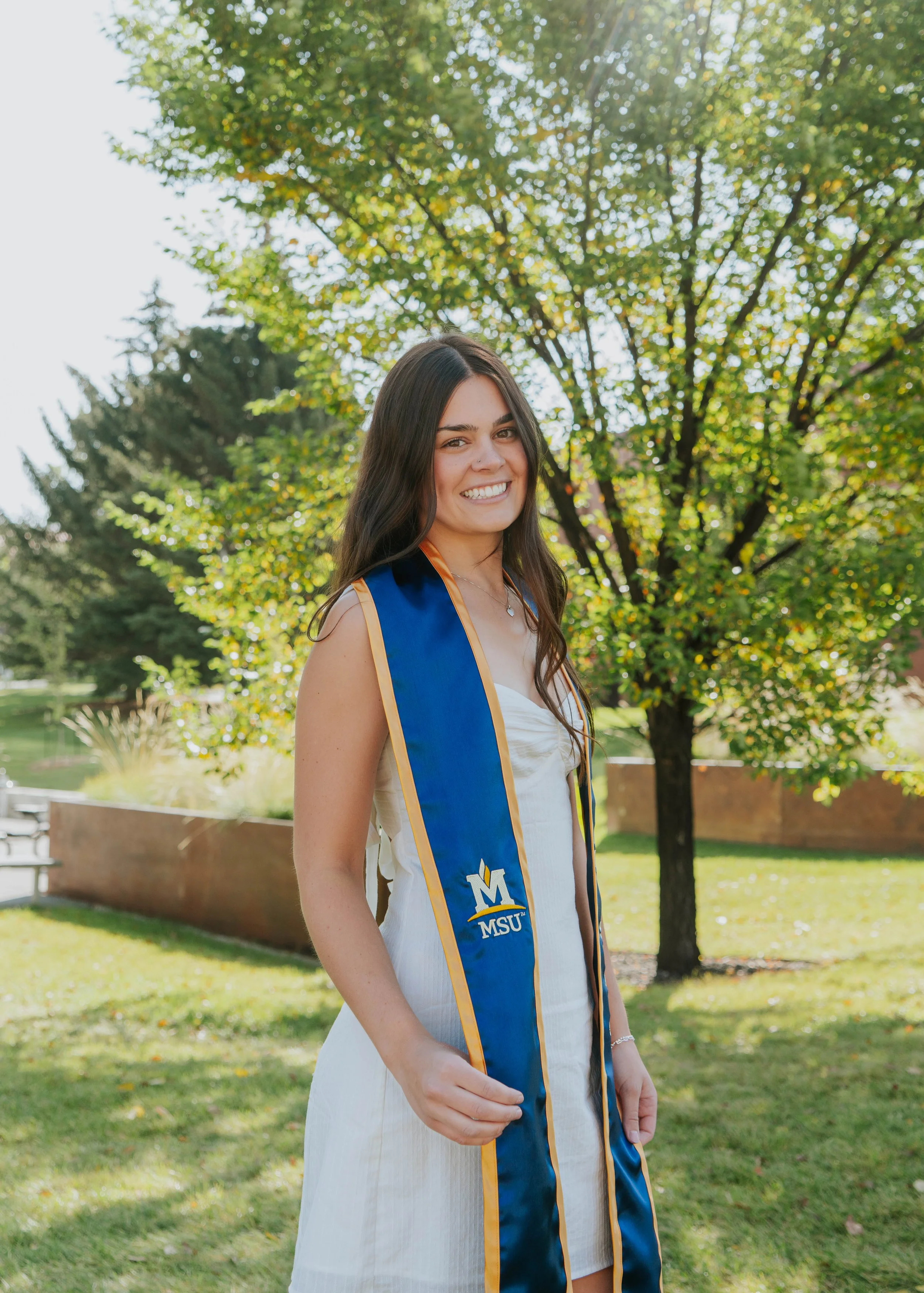 A young woman in a white dress wearing a blue MSU graduation stole, standing outdoors in front of green trees and smiling.