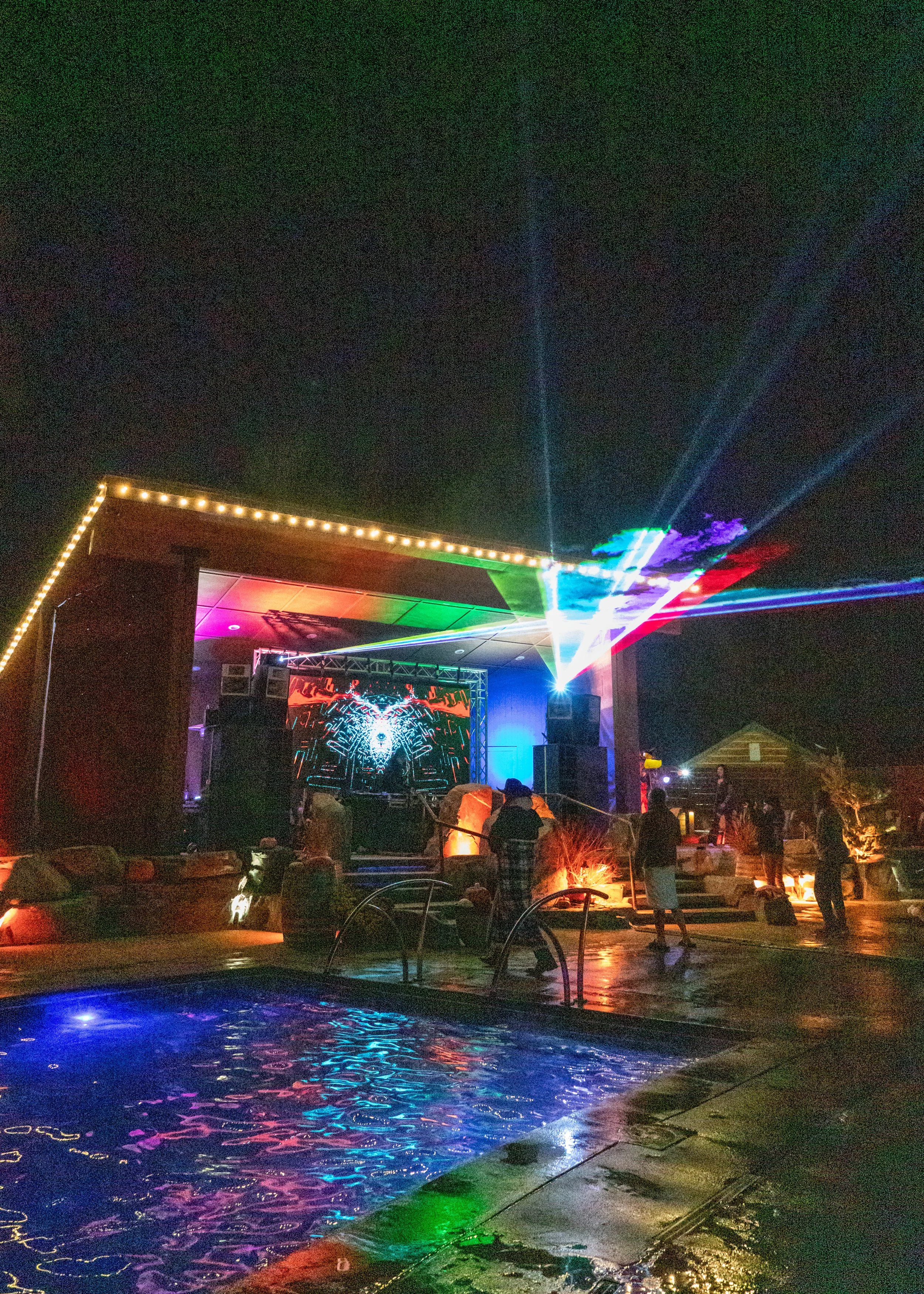 Nighttime outdoor party with a lit stage, colorful laser lights, and a swimming pool reflecting the lights.