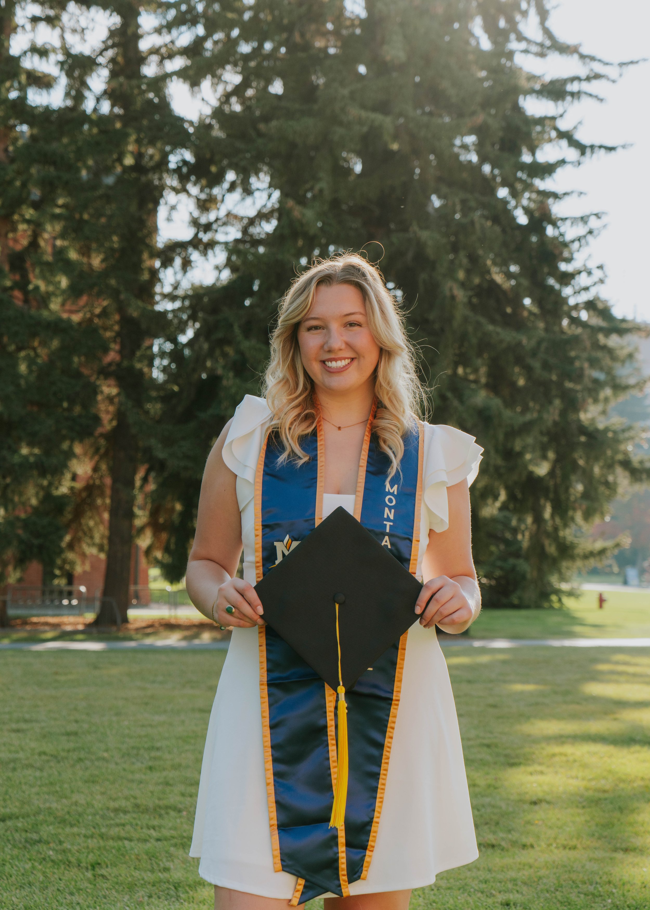A young woman in a white dress standing on grass during daytime, holding a graduation cap with a yellow tassel, wearing a blue sash with gold trim and the word "MONTANA" on it, smiling with trees and a building in the background.
