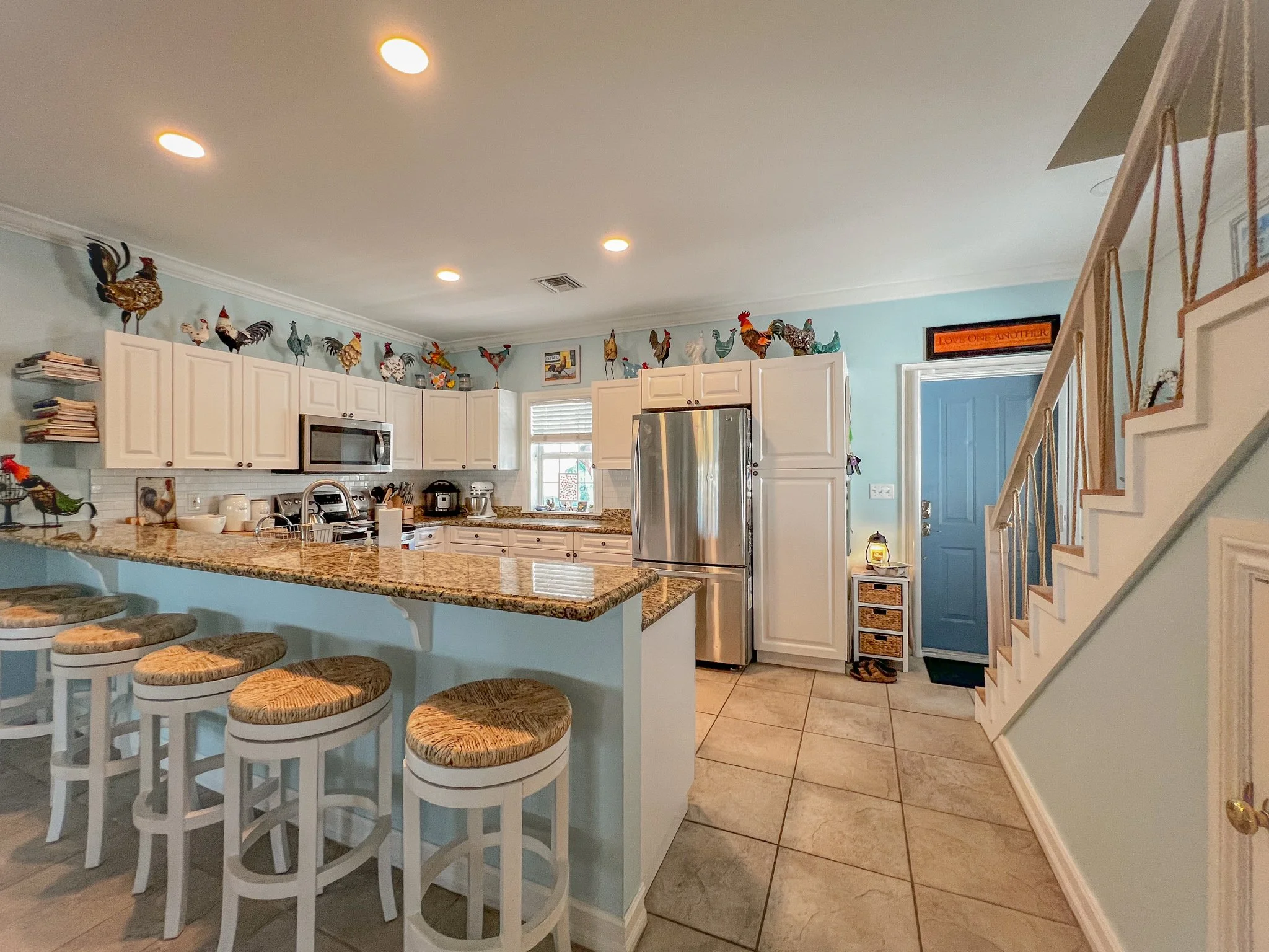Kitchen with white cabinets, beige granite countertops, stainless steel refrigerator, microwave, coffee maker, and decorative roosters on top of cabinets. Blue door, small wooden cabinet near door, and spiral staircase with wooden handrail.