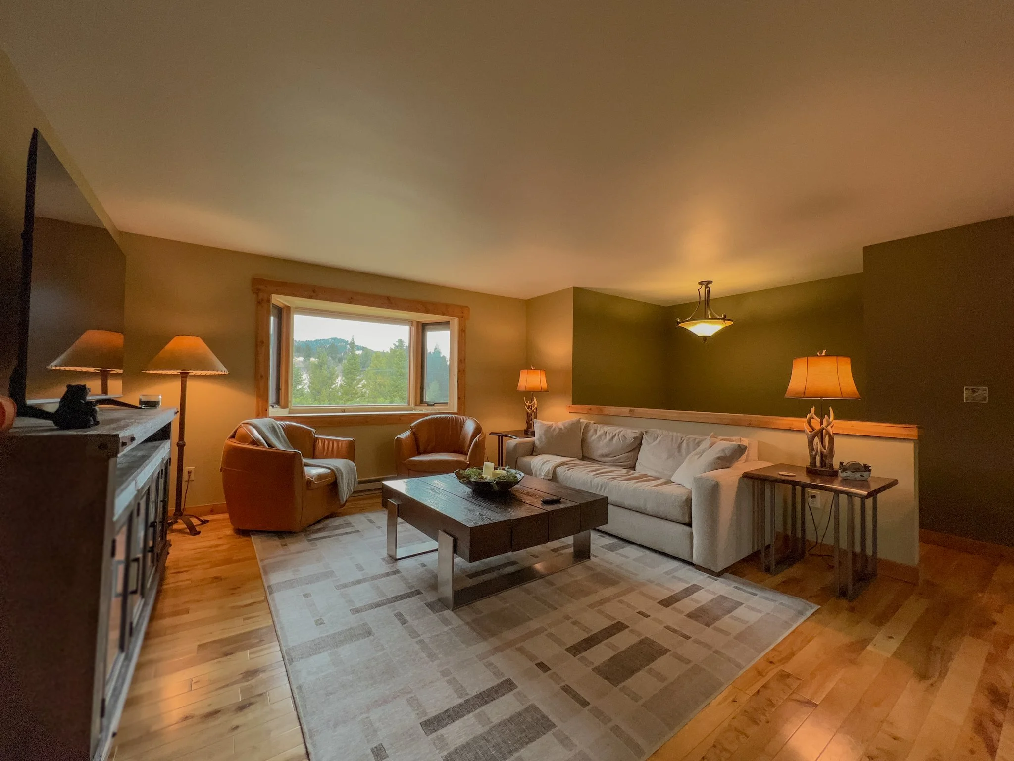 Living room with a large white sofa, two leather armchairs, a wooden coffee table, a patterned rug, side tables with lamps, a window showing a view of trees and hills, and a ceiling light fixture.