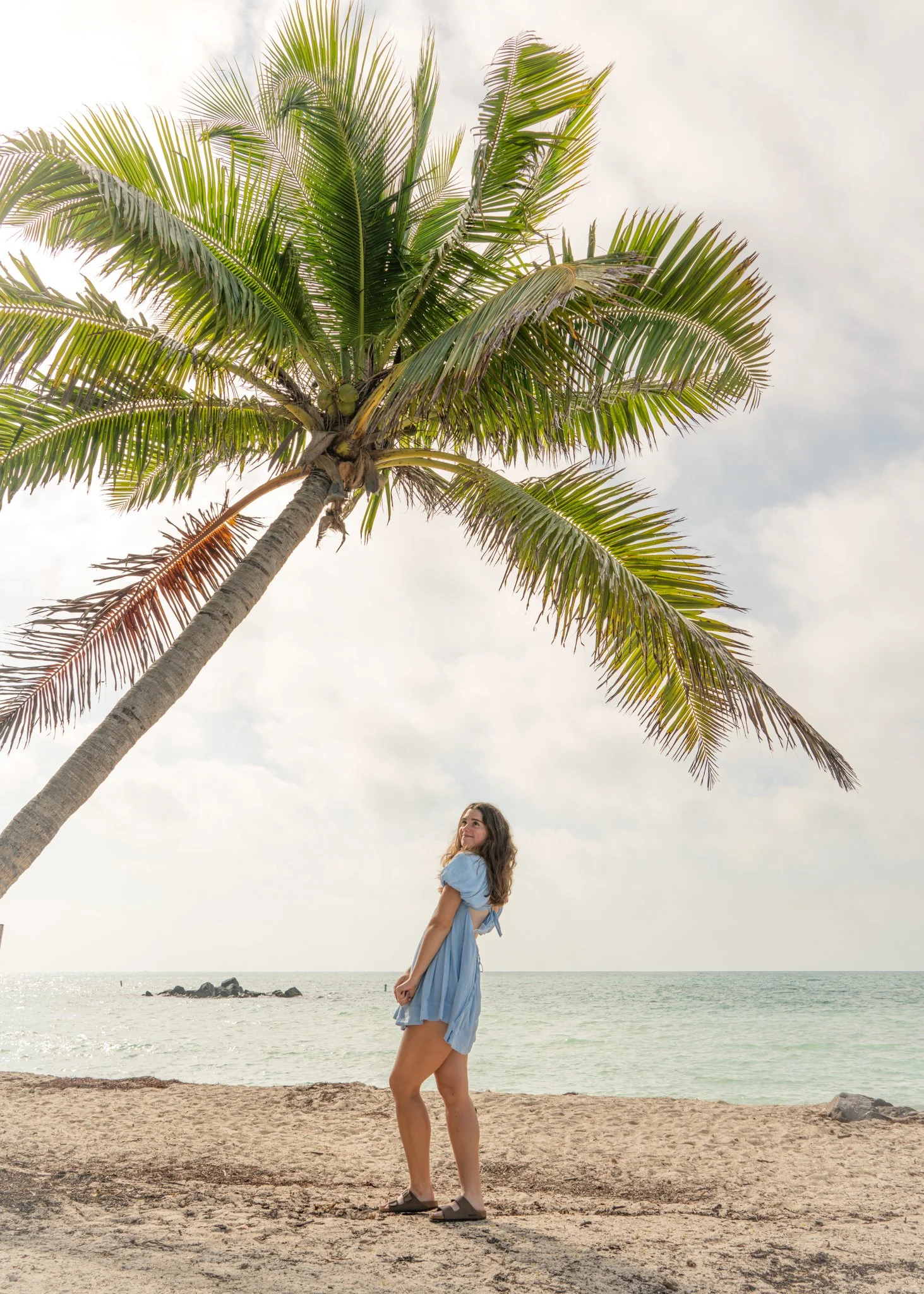 Girl standing on sandy beach under a large palm tree with overcast sky and ocean in the background.