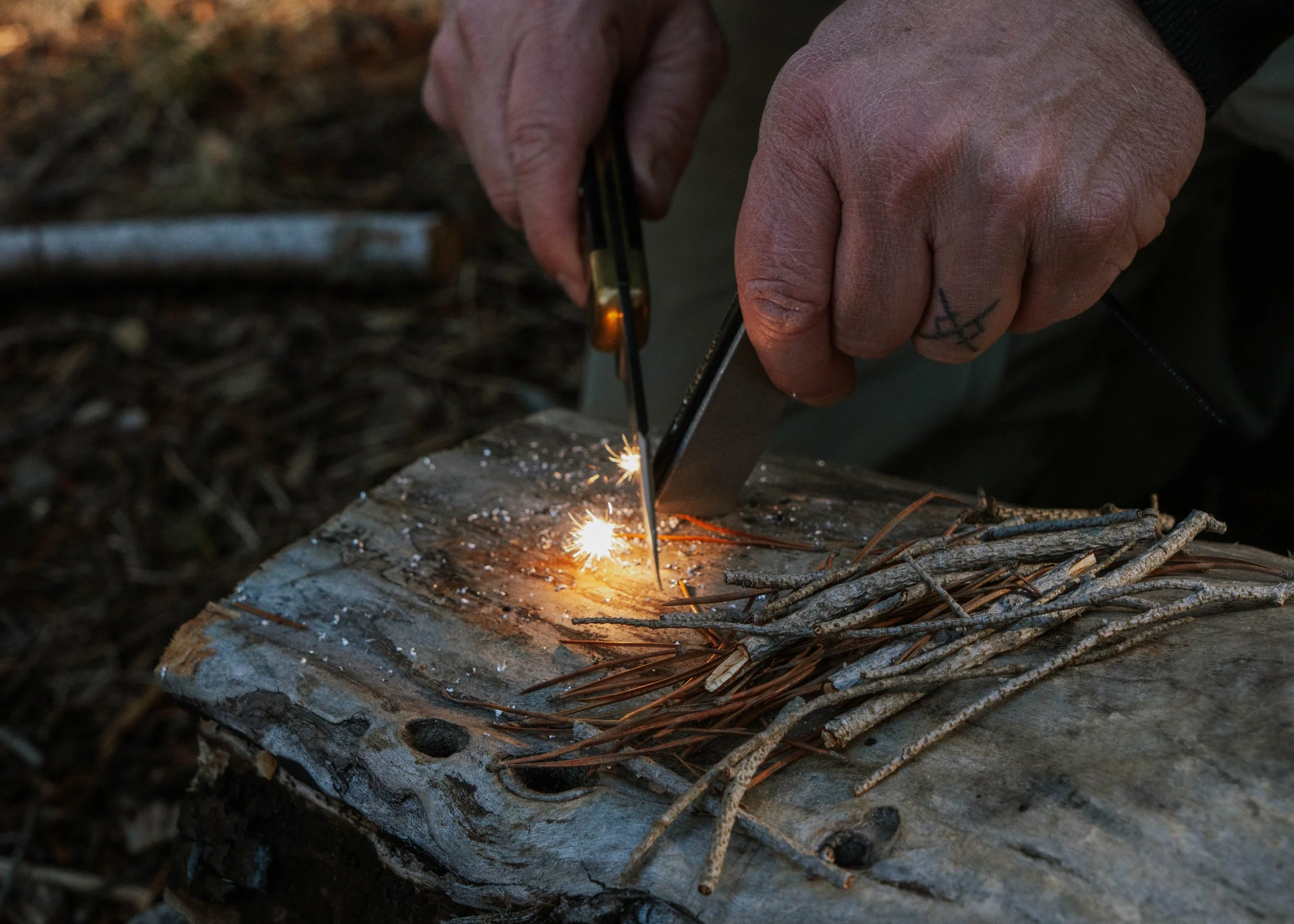 A person lighting kindling on a log with a spark from a fire starter. The person's hands are visible, with one hand holding a fire starter and the other using a striker. The kindling includes small twigs placed on the log.