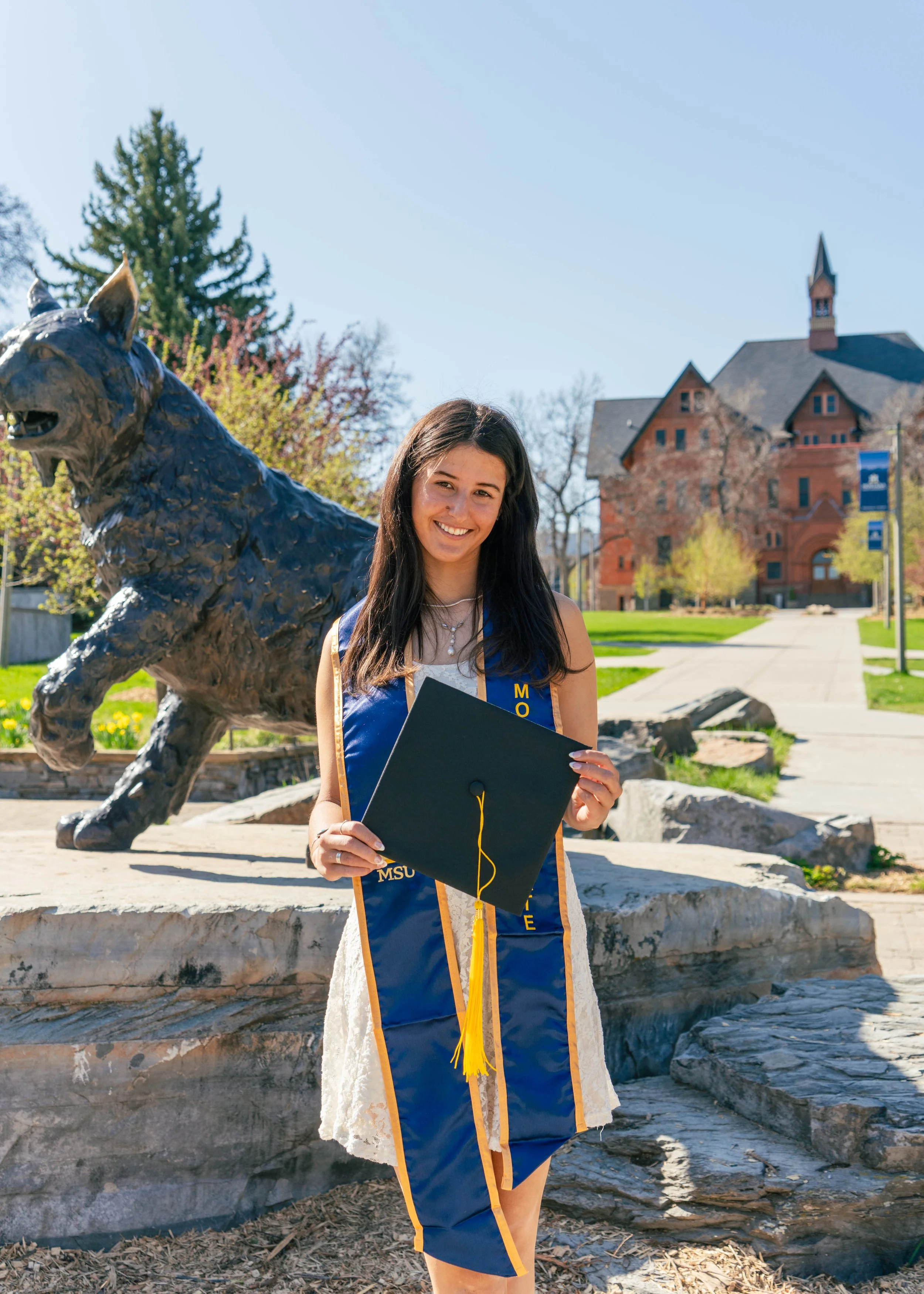 Smiling young woman in graduation gown and cap holding diploma outside near a statue of a wildcat on a university campus with historic brick building in the background.