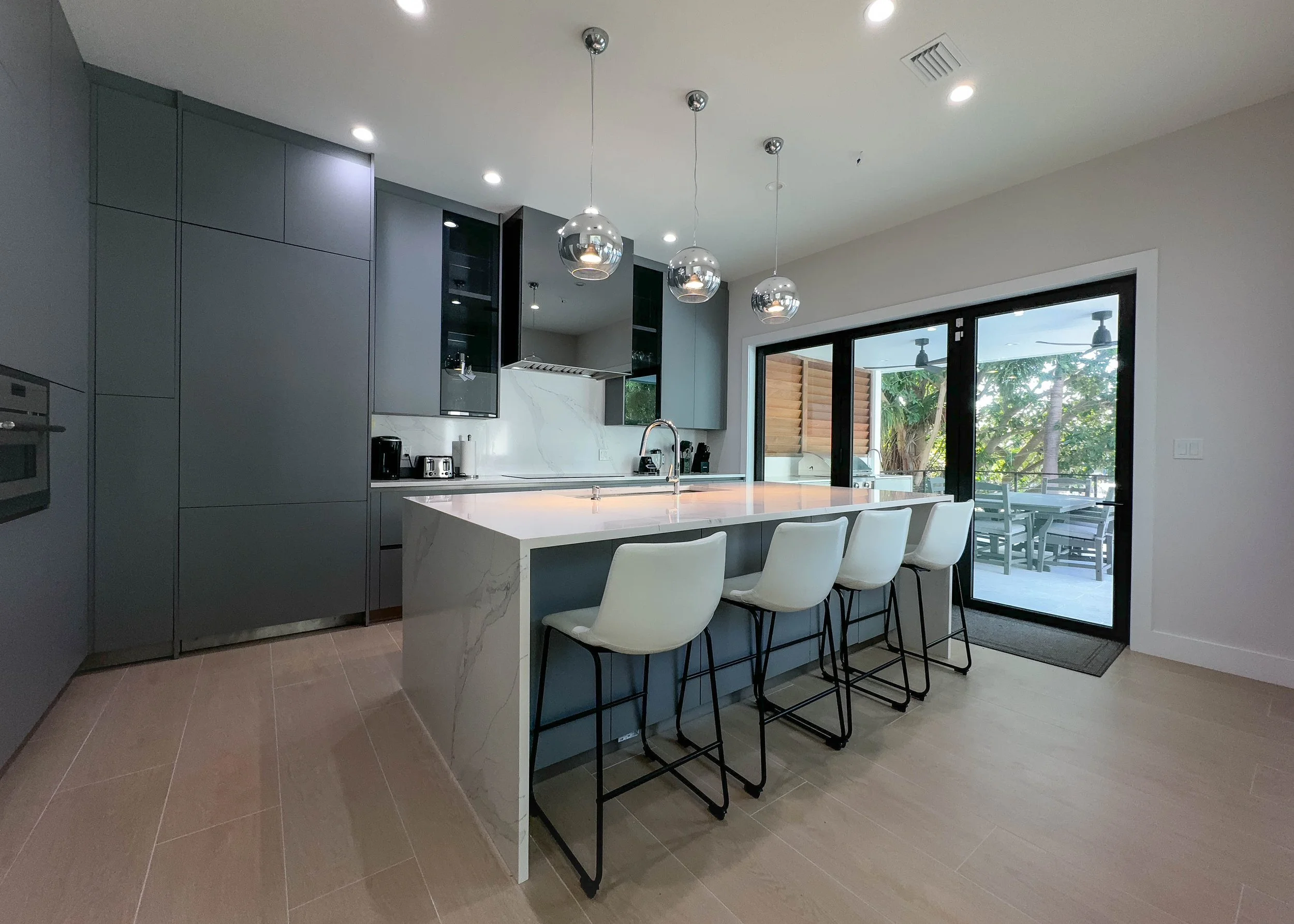 Modern kitchen with gray cabinetry, white marble island, four white barstools, and sliding glass doors leading to an outdoor patio.