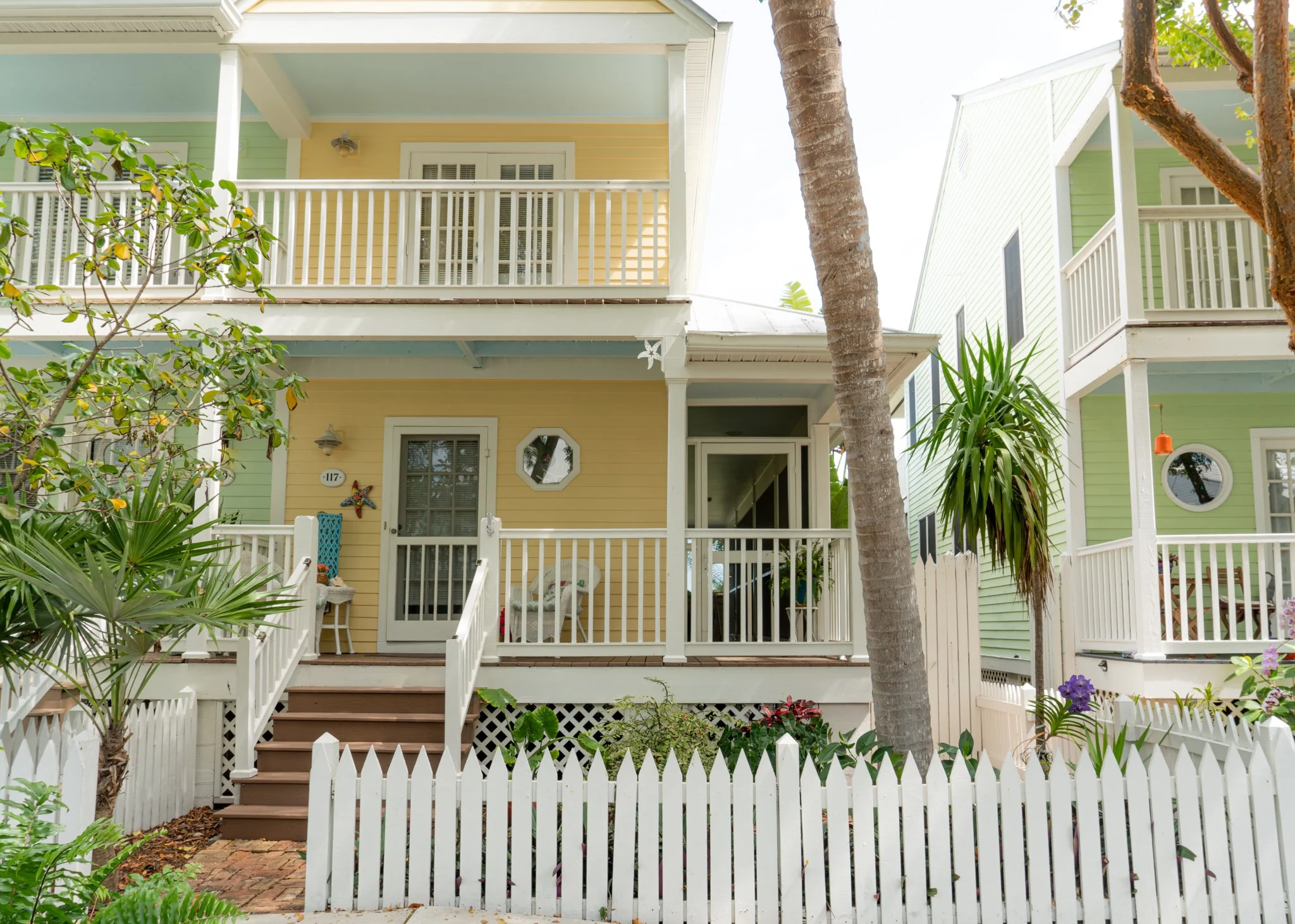 Colorful two-story house with a yellow facade, white trim, and a porch, surrounded by lush tropical plants and a white picket fence.