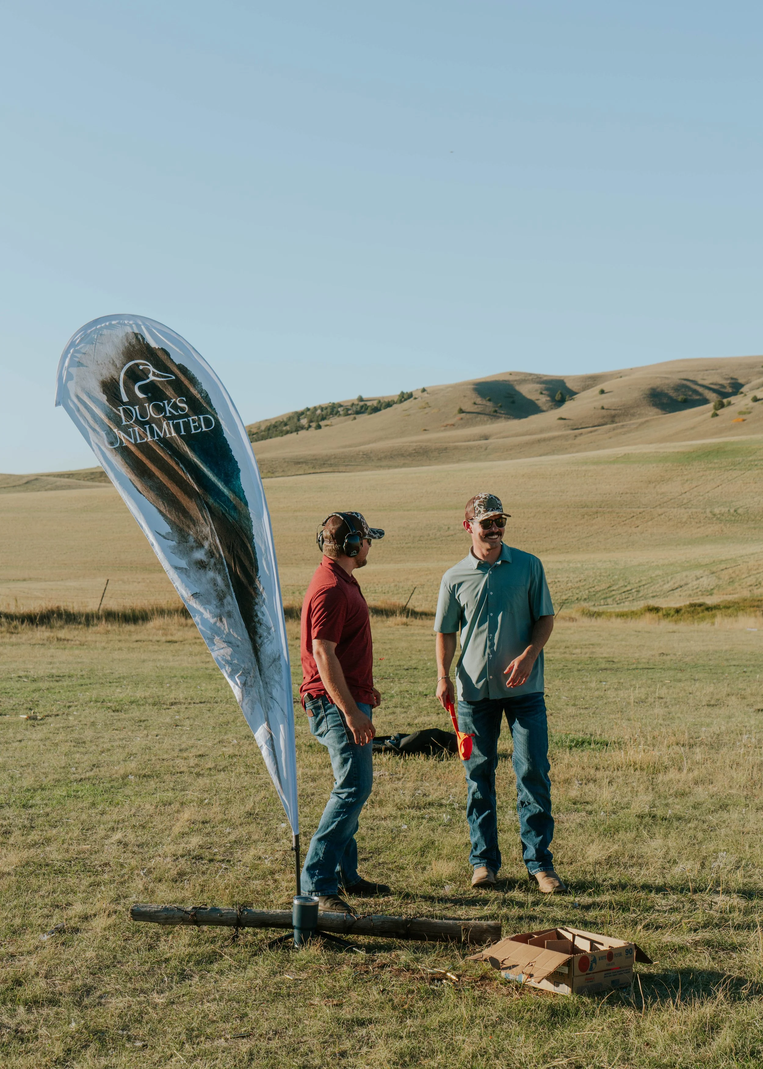 Two men standing outdoors in a grassy field with hills in the background, one holding a red object and there is a flag that says 'Ducks Unlimited,' with some cardboard boxes on the ground.