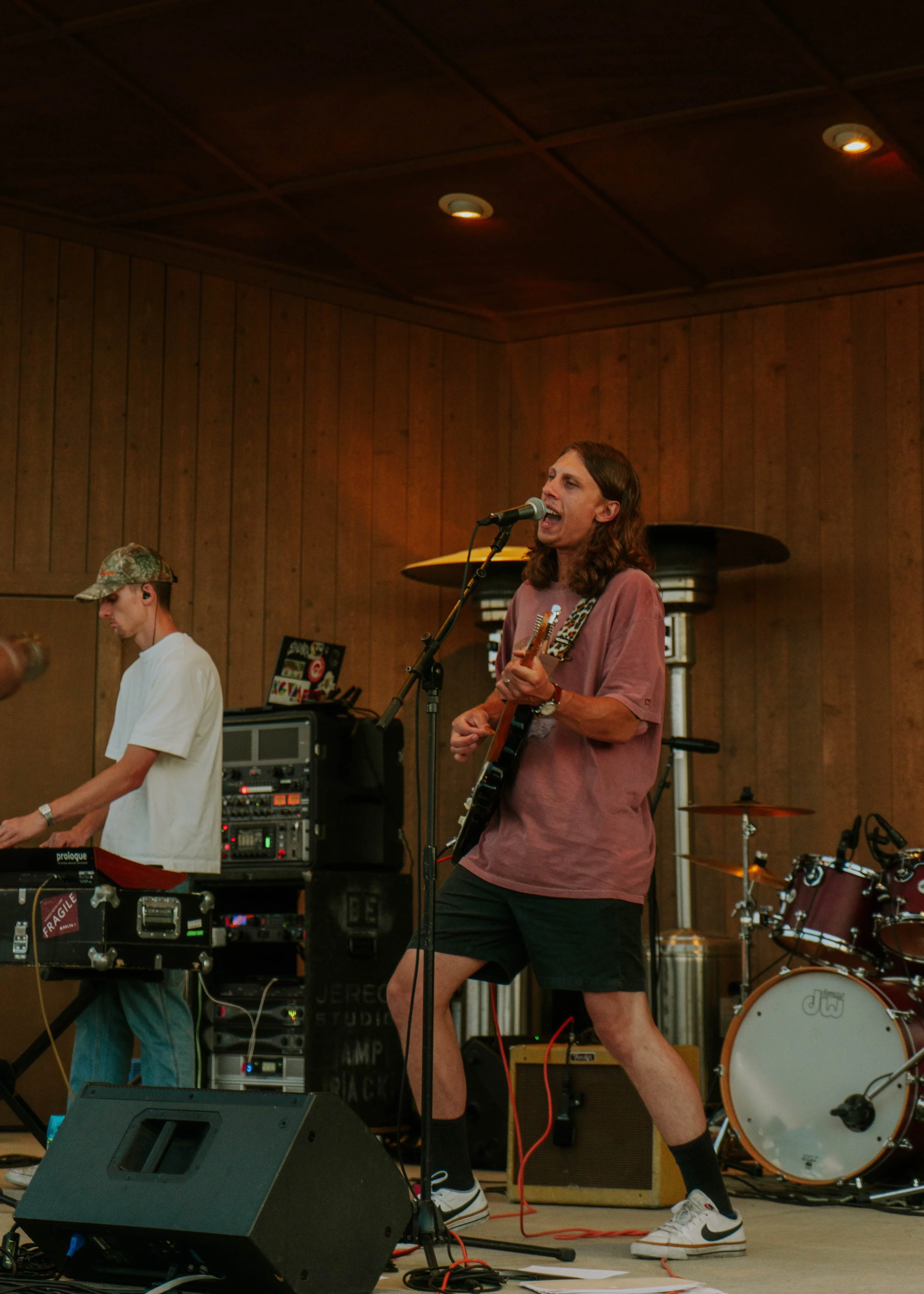 A male musician with long hair singing into a microphone, playing an electric guitar, wearing a pink T-shirt, black shorts, and Nike sneakers, performing on stage with a keyboard player and a drum set in the background.