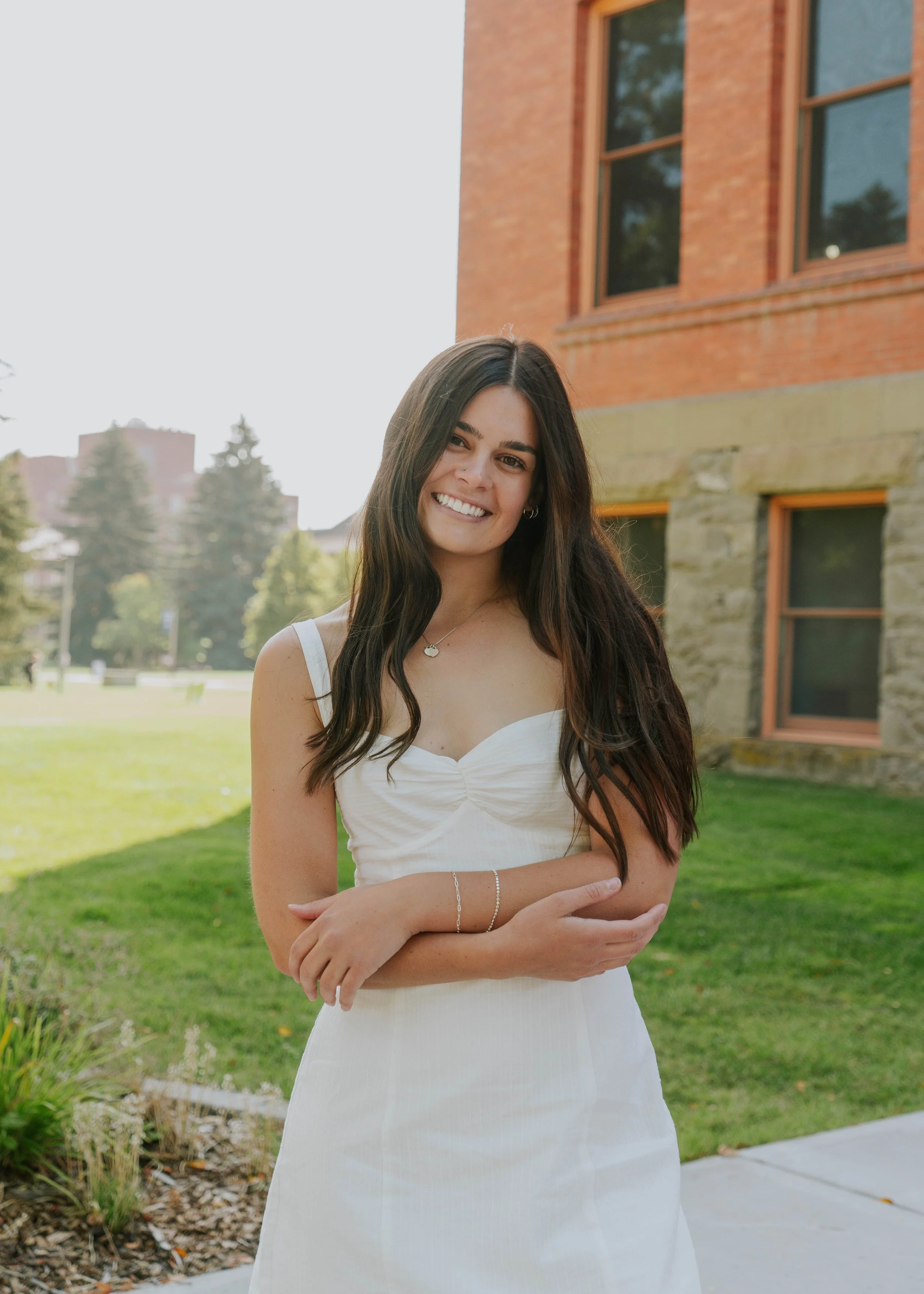 A smiling young woman with long brown hair, wearing a white dress, standing outside in front of a brick and stone building with green grass and trees in the background.