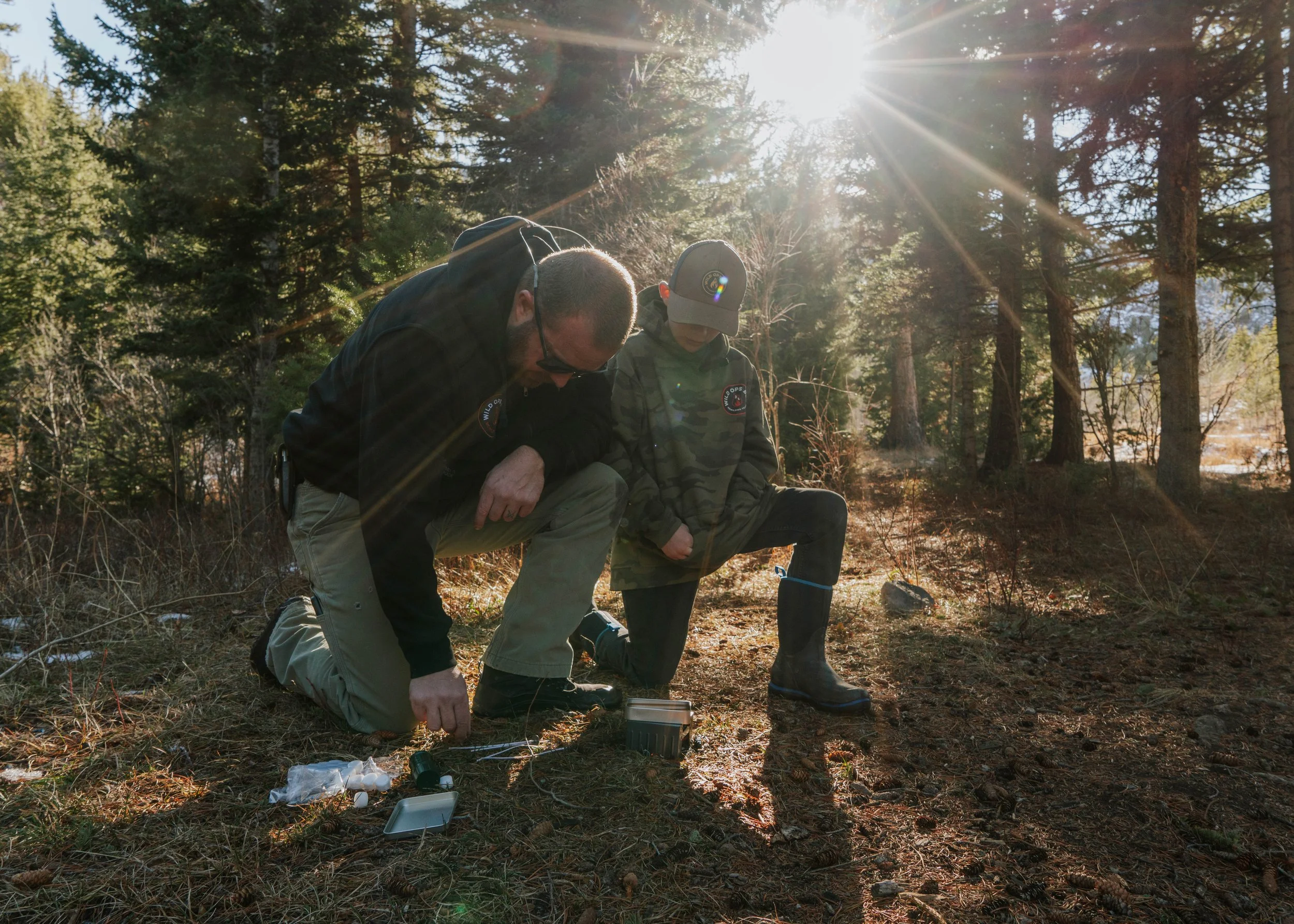 Two men kneeling on the ground in a wooded area, working with scientific equipment, with sunlight shining through trees.