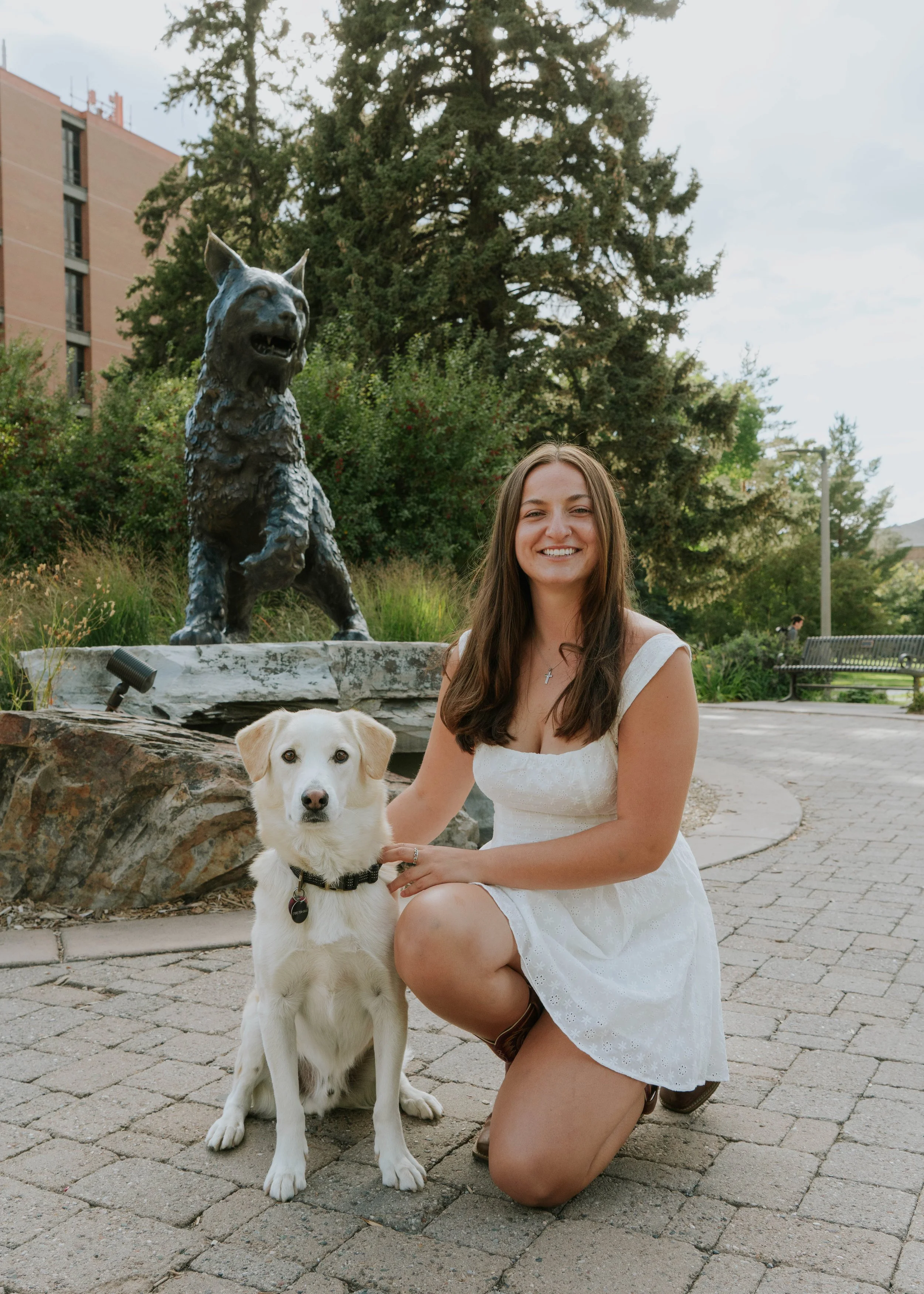 A woman in a white dress kneeling next to a white dog with a black collar, outdoors in front of a statue of a wolf on a stone base, with trees and a bench in the background.