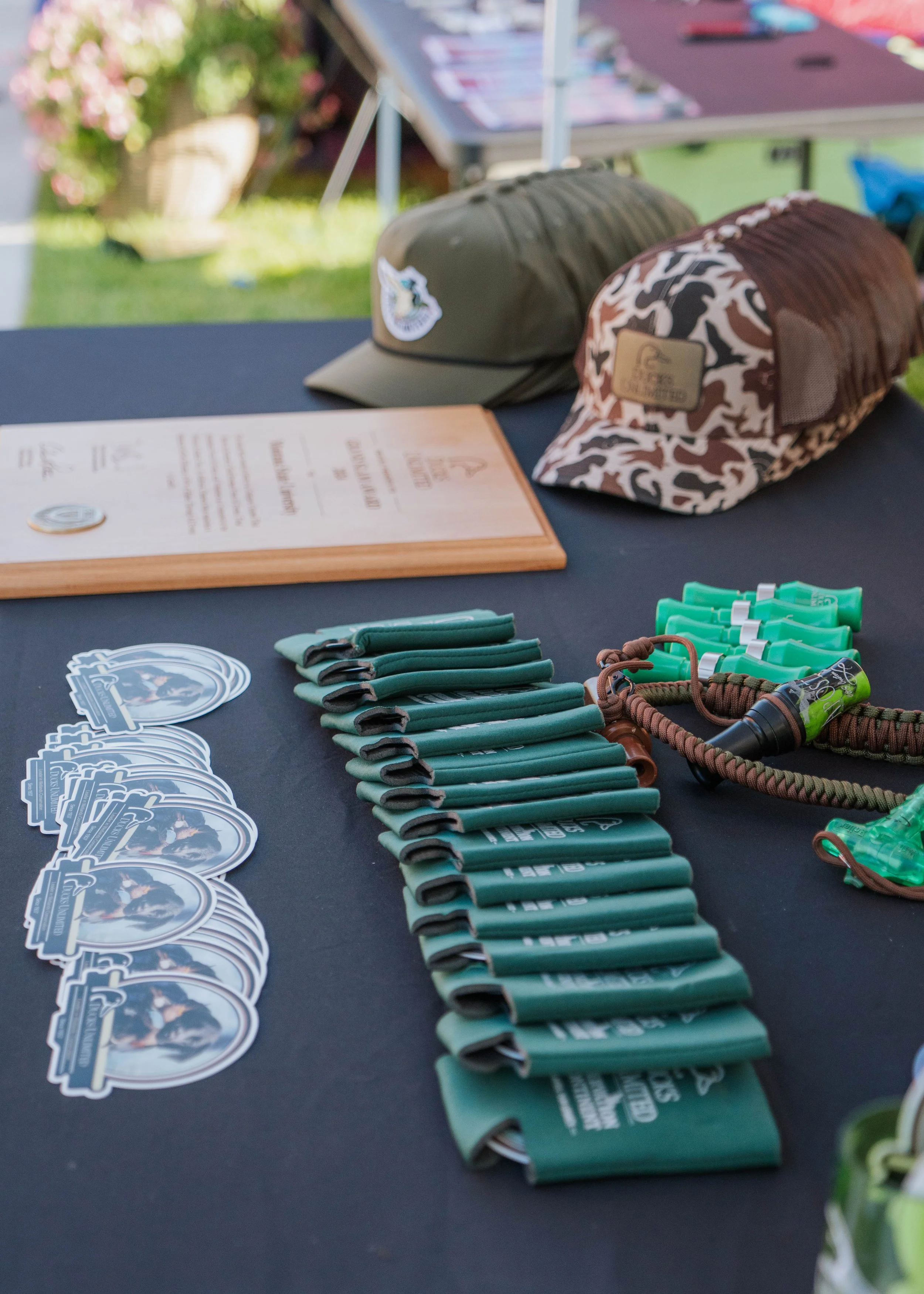 Table with green hats, stickers, and lanyards displayed outdoors, with hats featuring outdoor logos, and a menu in the background.