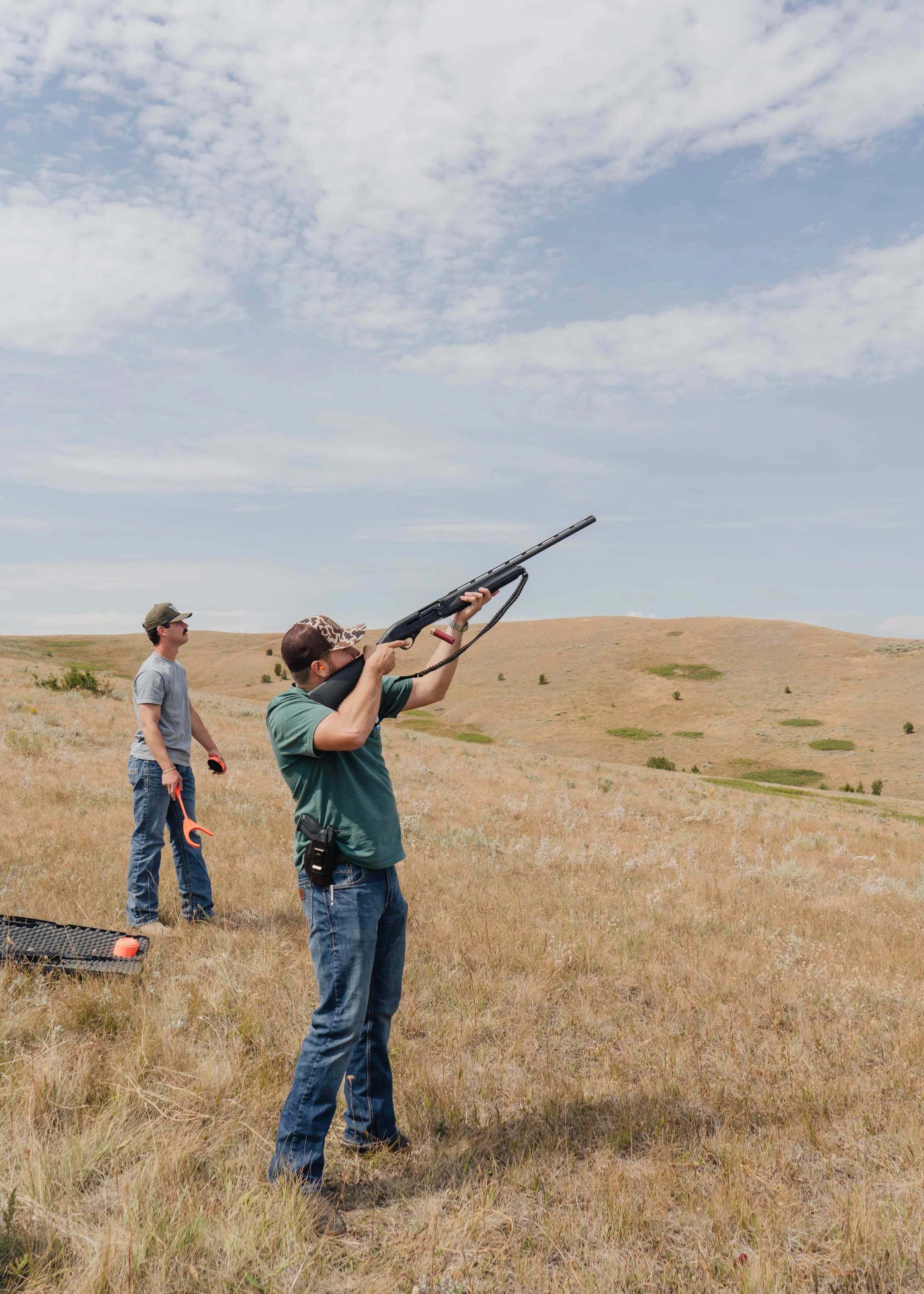 Two men outdoors in a grassy field, one aiming a shotgun and the other observing, under a partly cloudy sky.