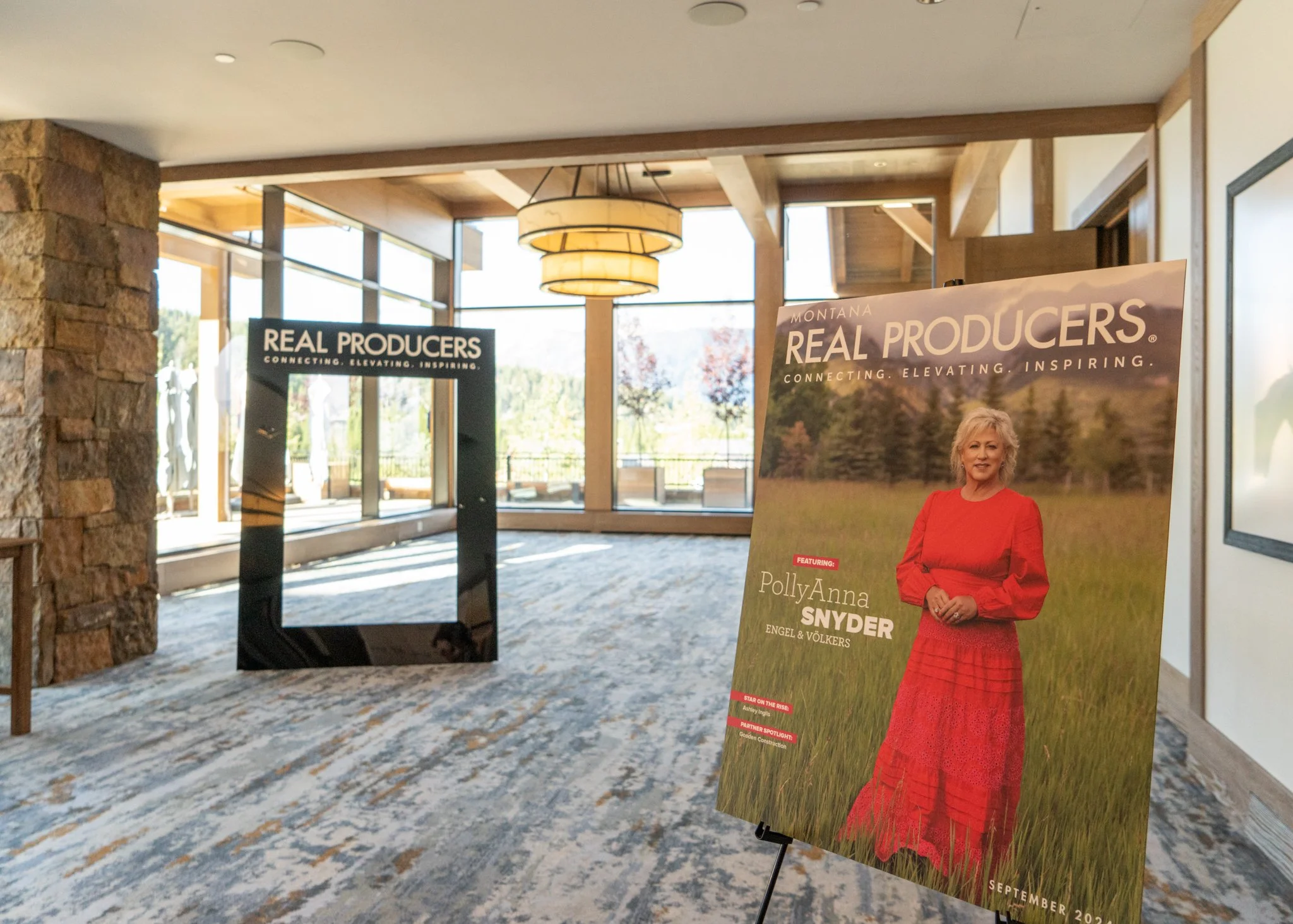 Interior of a conference or event space with promotional displays including a large poster featuring PollyAnna Snyder in a red dress standing in a field, and a black standee with the text 'Real Producers'.