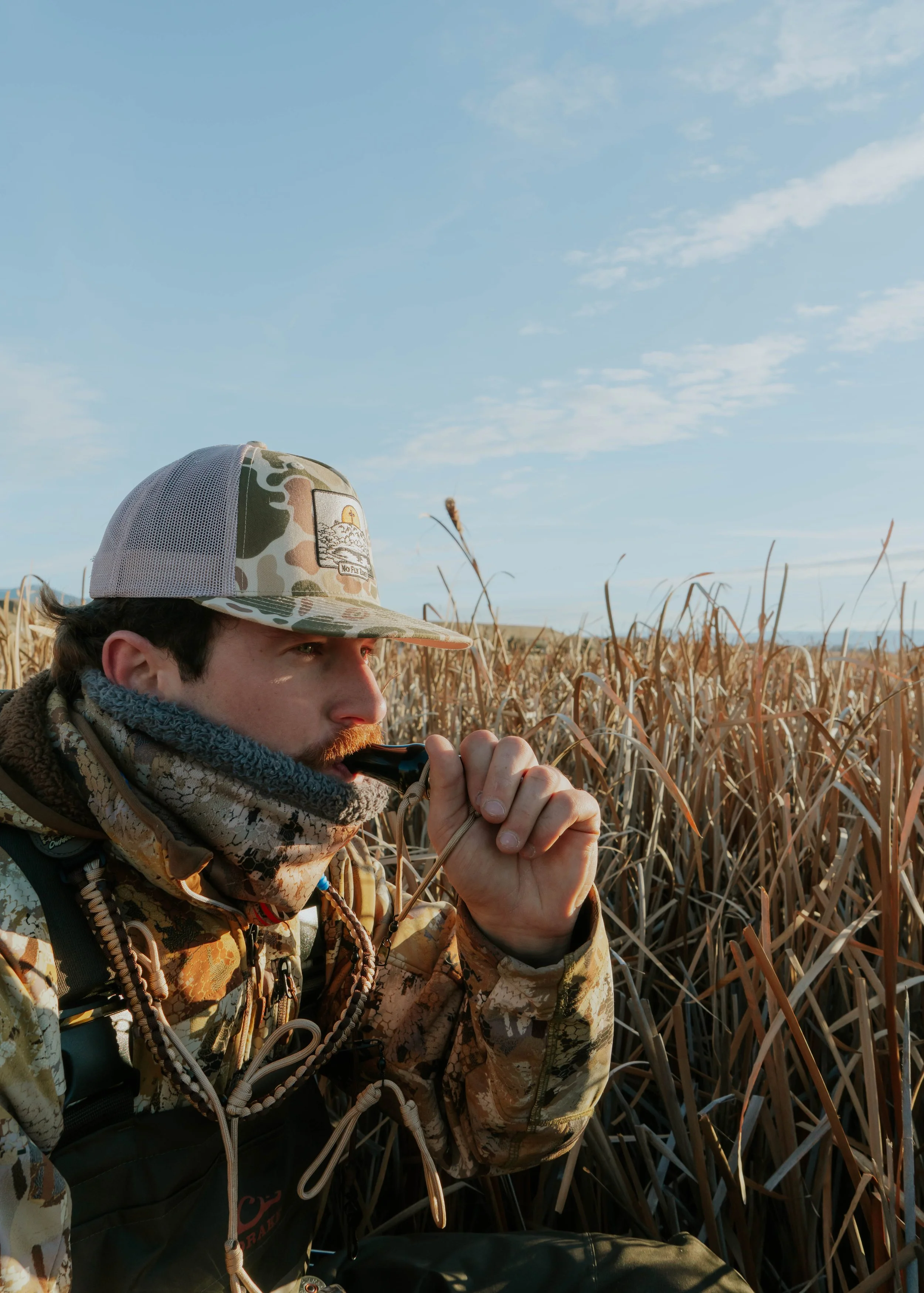A man in camouflage outdoor gear and a camo hat is sitting among tall, dry grass, holding a small pipe to his lips, in a field during daylight with a clear blue sky.