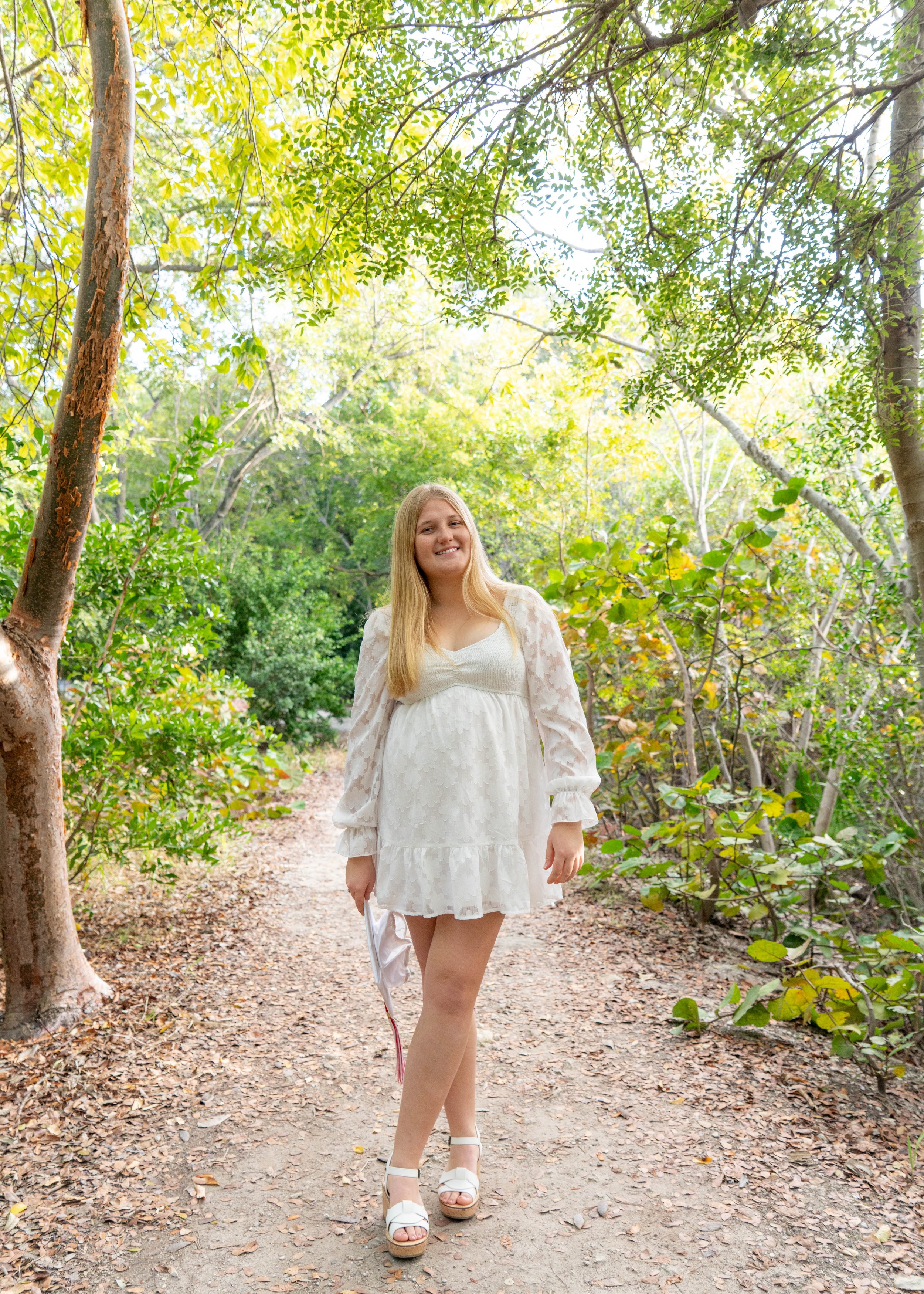 A young woman with long blonde hair wearing a white dress with long sleeves and platform sandals, standing on a wooded trail surrounded by green trees and foliage, holding a white hat in her left hand, smiling at the camera.