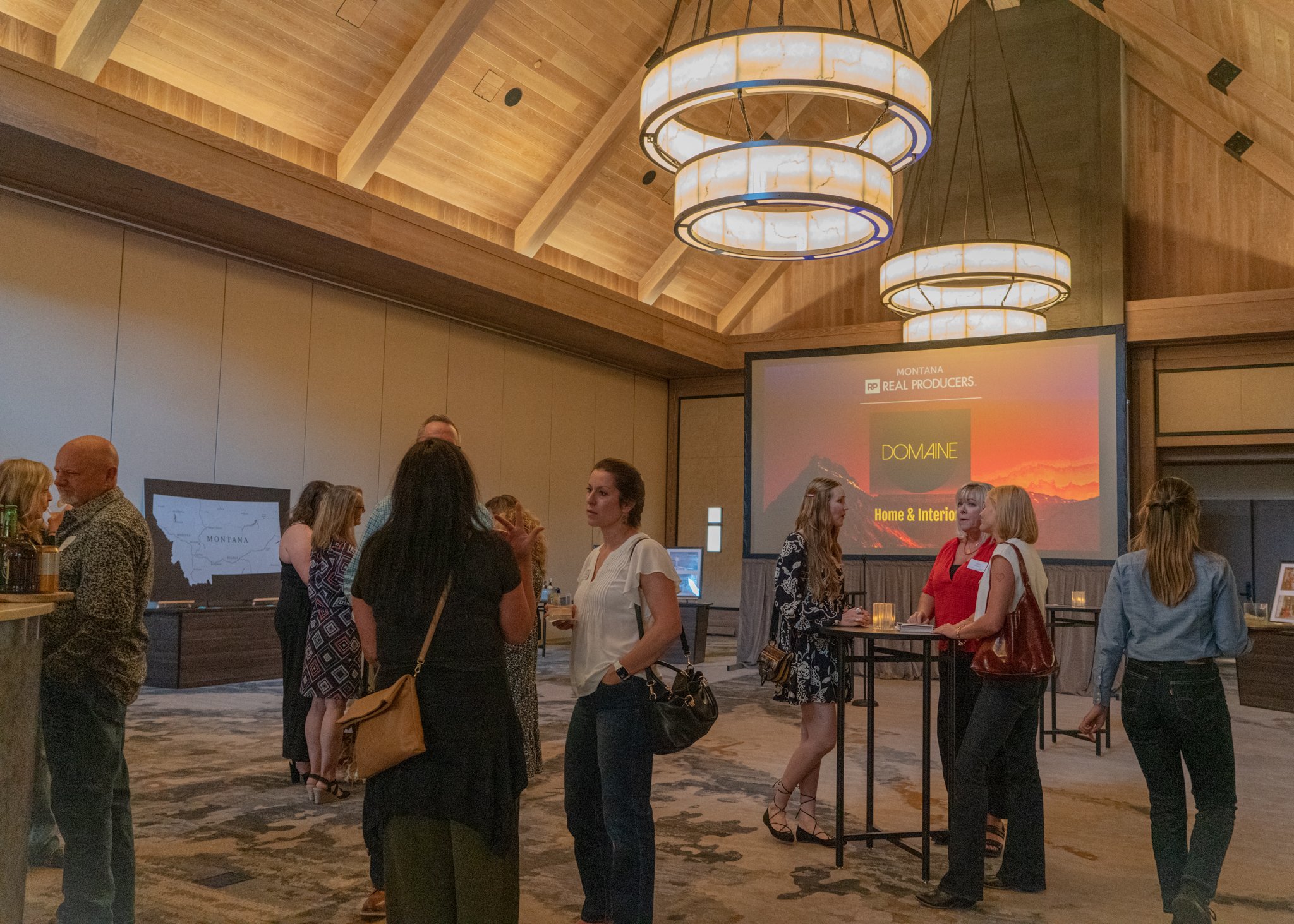 People attending a conference or business event in a spacious room with wooden ceiling, chandeliers, and a large screen displaying 'Montana Real Producers' and 'Domaine Home & Interior', with some individuals engaged in conversations.