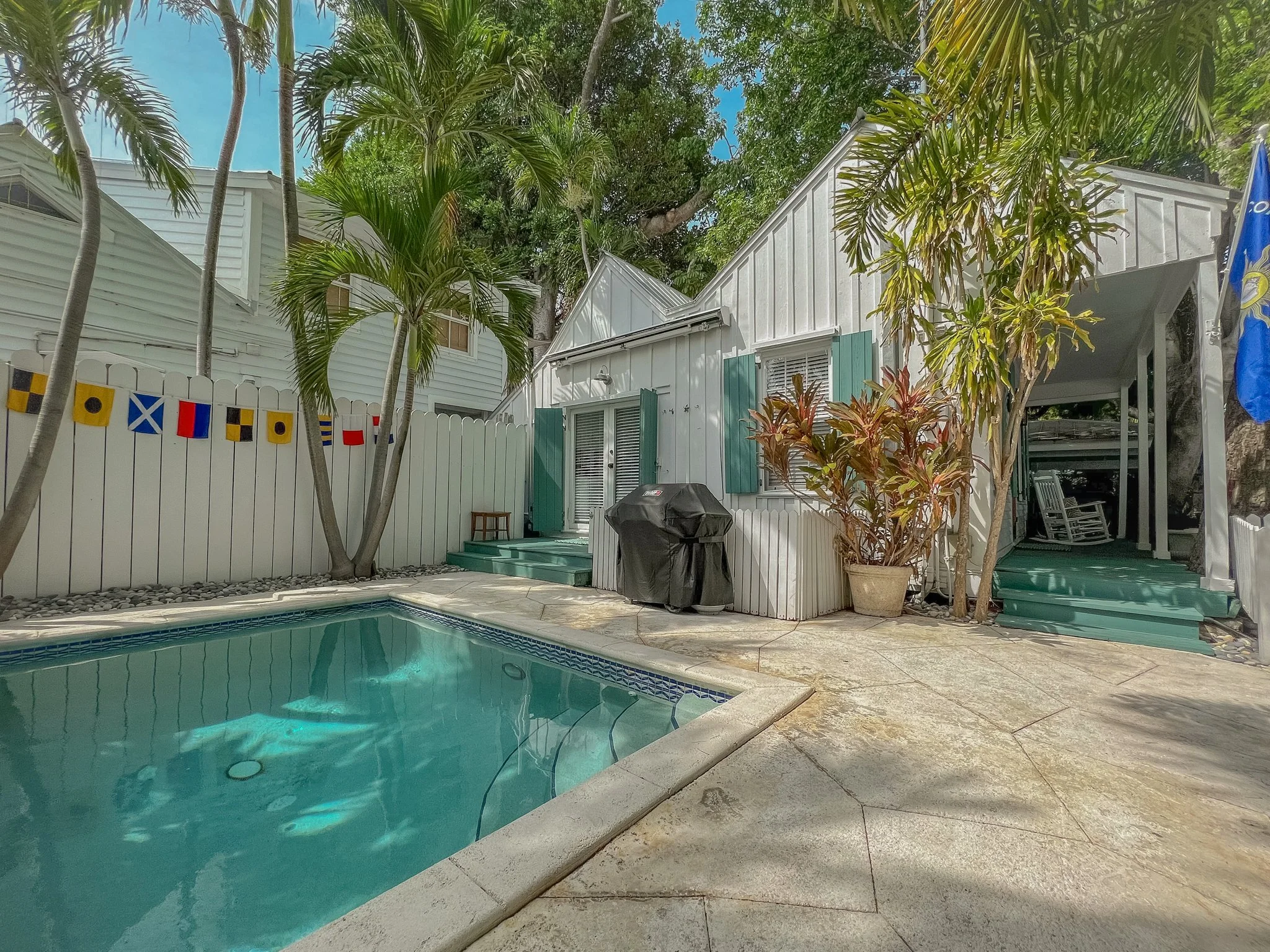 Backyard of a house with a small swimming pool, tropical plants, a barbecue grill, and a small deck area with chairs.