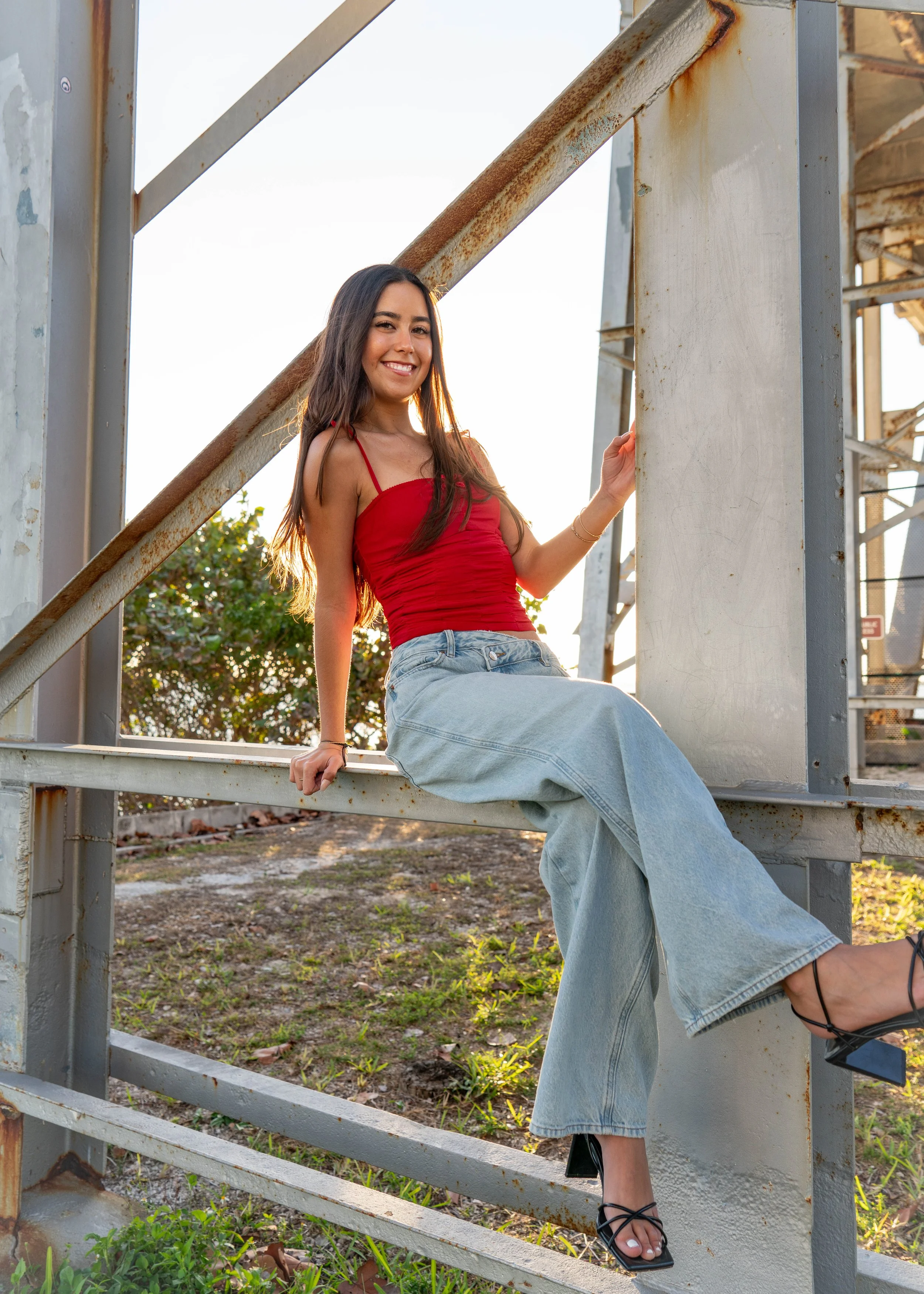 A young woman sits on a metal railing outdoors, wearing a red spaghetti strap top, light blue wide-leg jeans, and black high-heeled sandals, smiling at the camera with sunlight behind her.