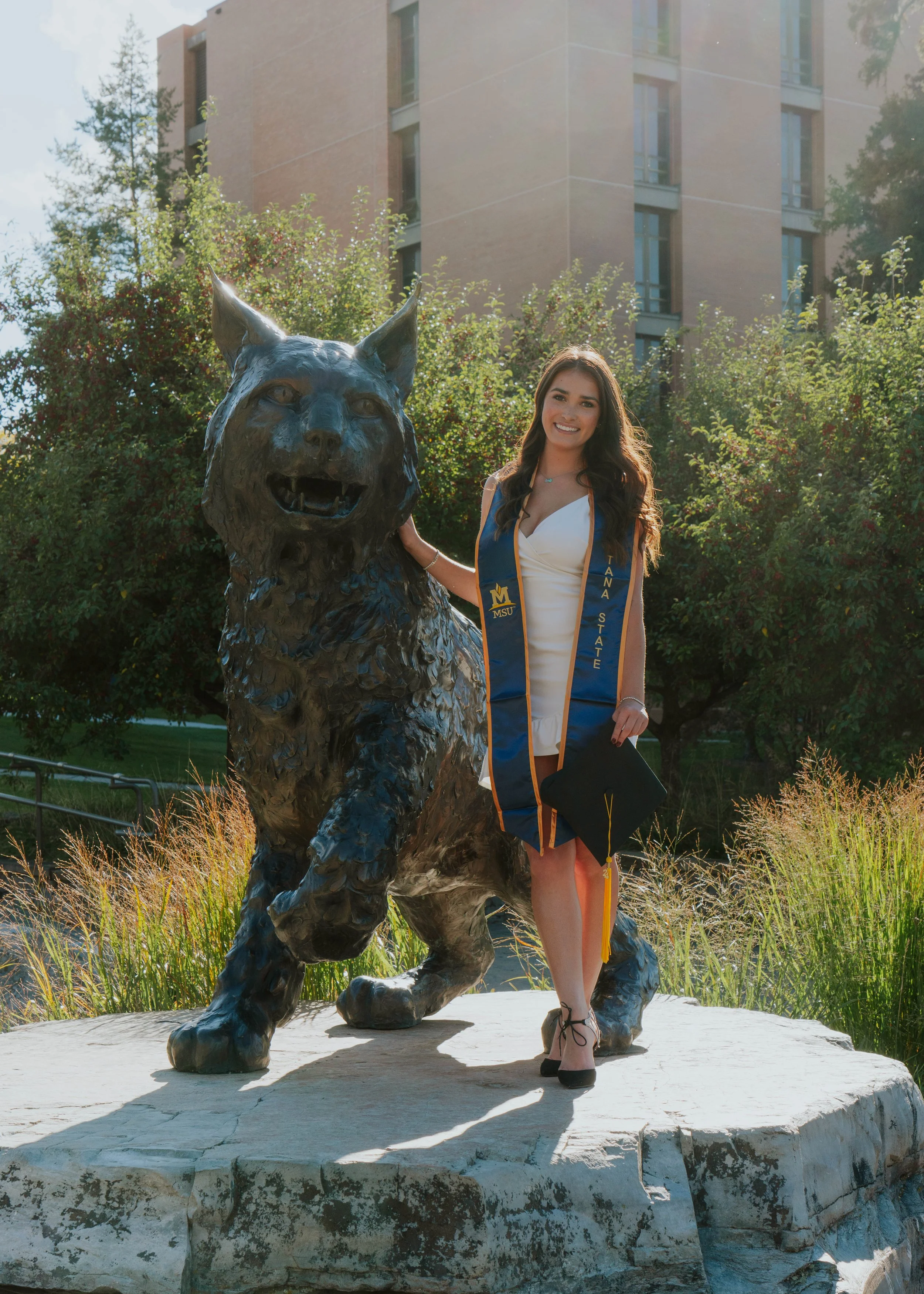 A young woman in a white dress and black high heels posing with a bronze statue of a cougar on a university campus. She is wearing a blue and gold graduation stole that reads "TANA STATE" and holds a cap in her hand, smiling at the camera. Trees and a building are in the background.