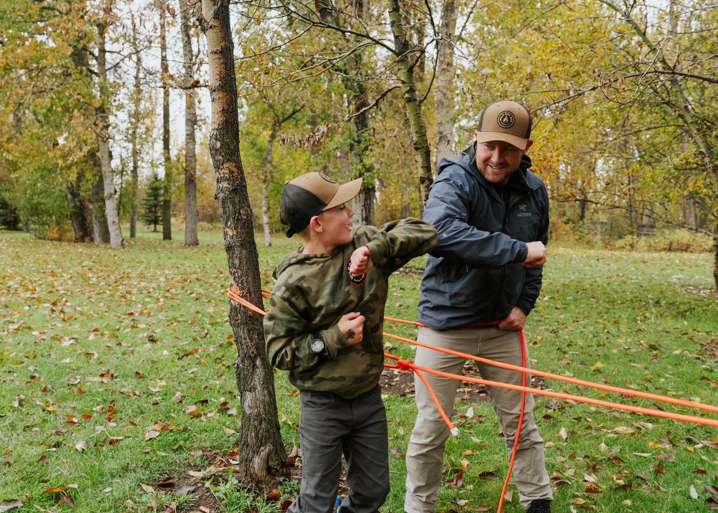 Two males, one younger and one adult, engage in a playful arm wrestling outside among trees with fall foliage, smiling and wearing hats and jackets.