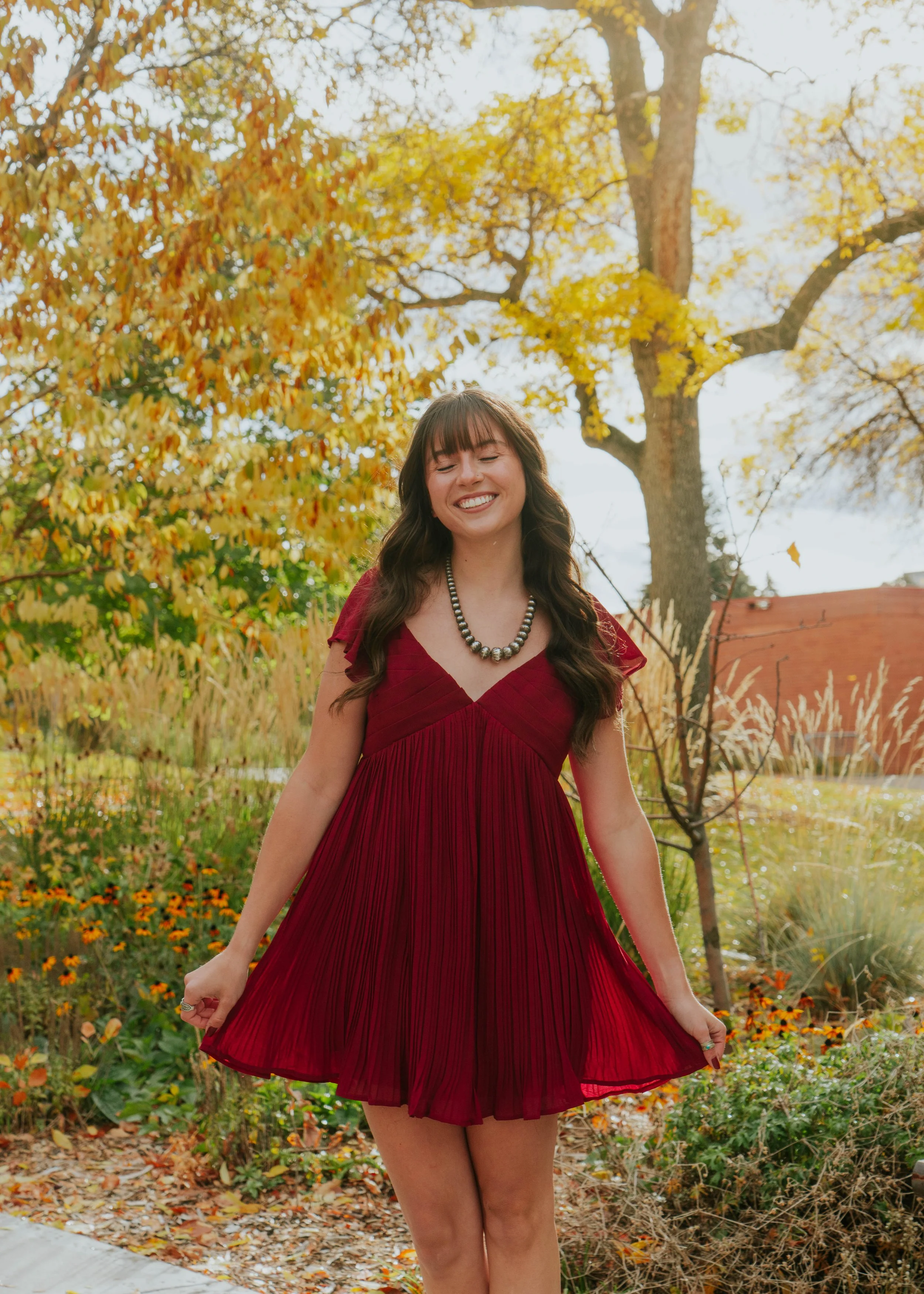 A young woman with long dark hair wearing a red dress and a pearl necklace, standing outdoors in a park during autumn with colorful trees and flowers in the background, smiling with eyes closed.