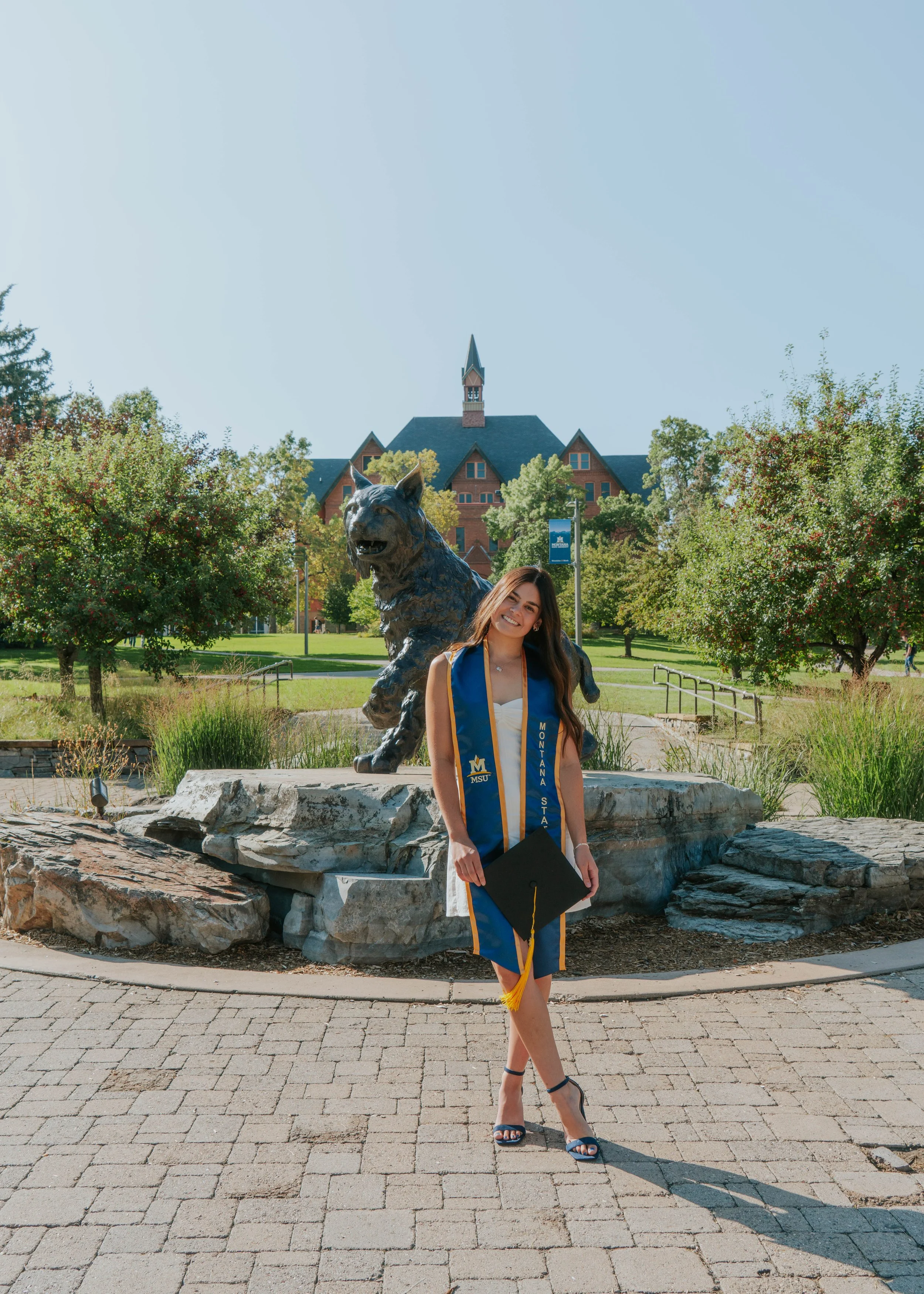 A young woman in a graduation gown and stole stands in front of a statue of a tiger on a college campus, holding a diploma and smiling.