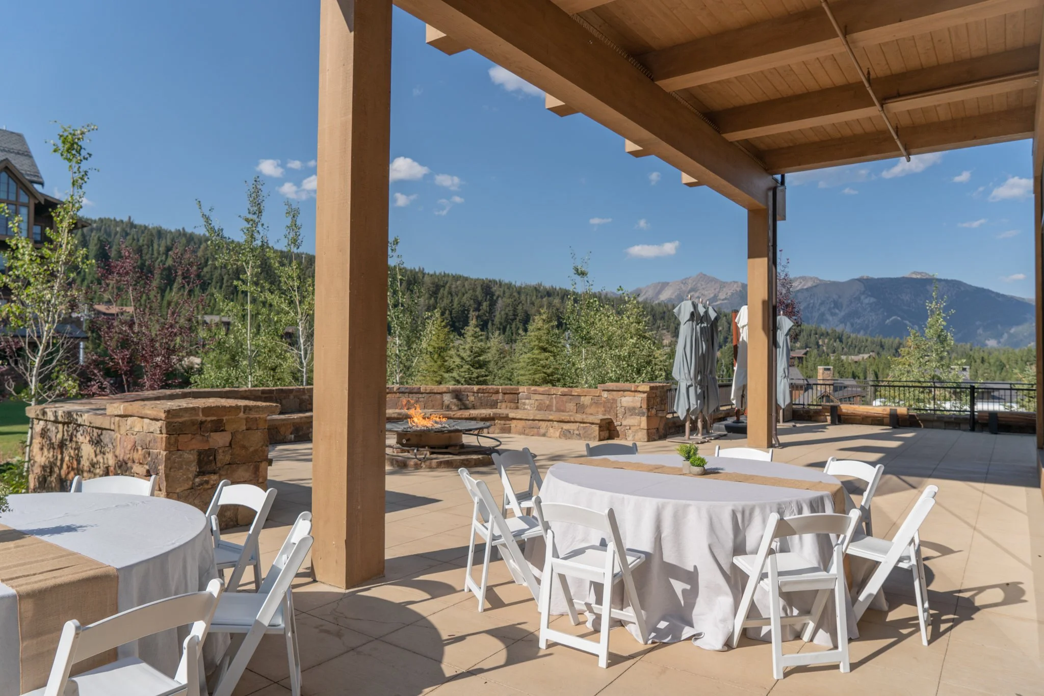 Outdoor patio with round tables covered with white tablecloths, chairs, a fire pit, and mountain view in the background.