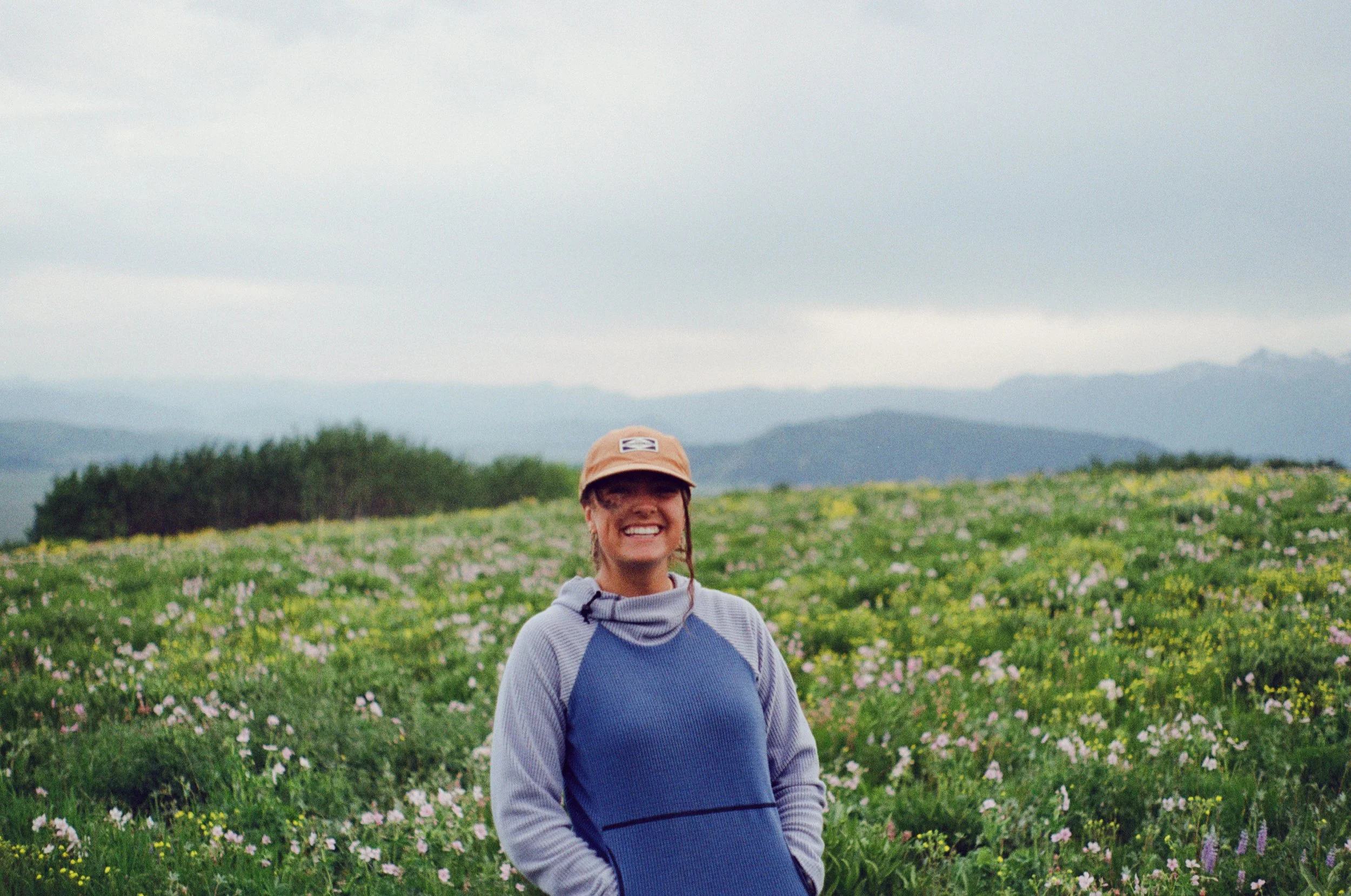 Smiling woman in a blue and gray jacket and tan cap standing in a field of wildflowers with mountains in the background on a cloudy day.
