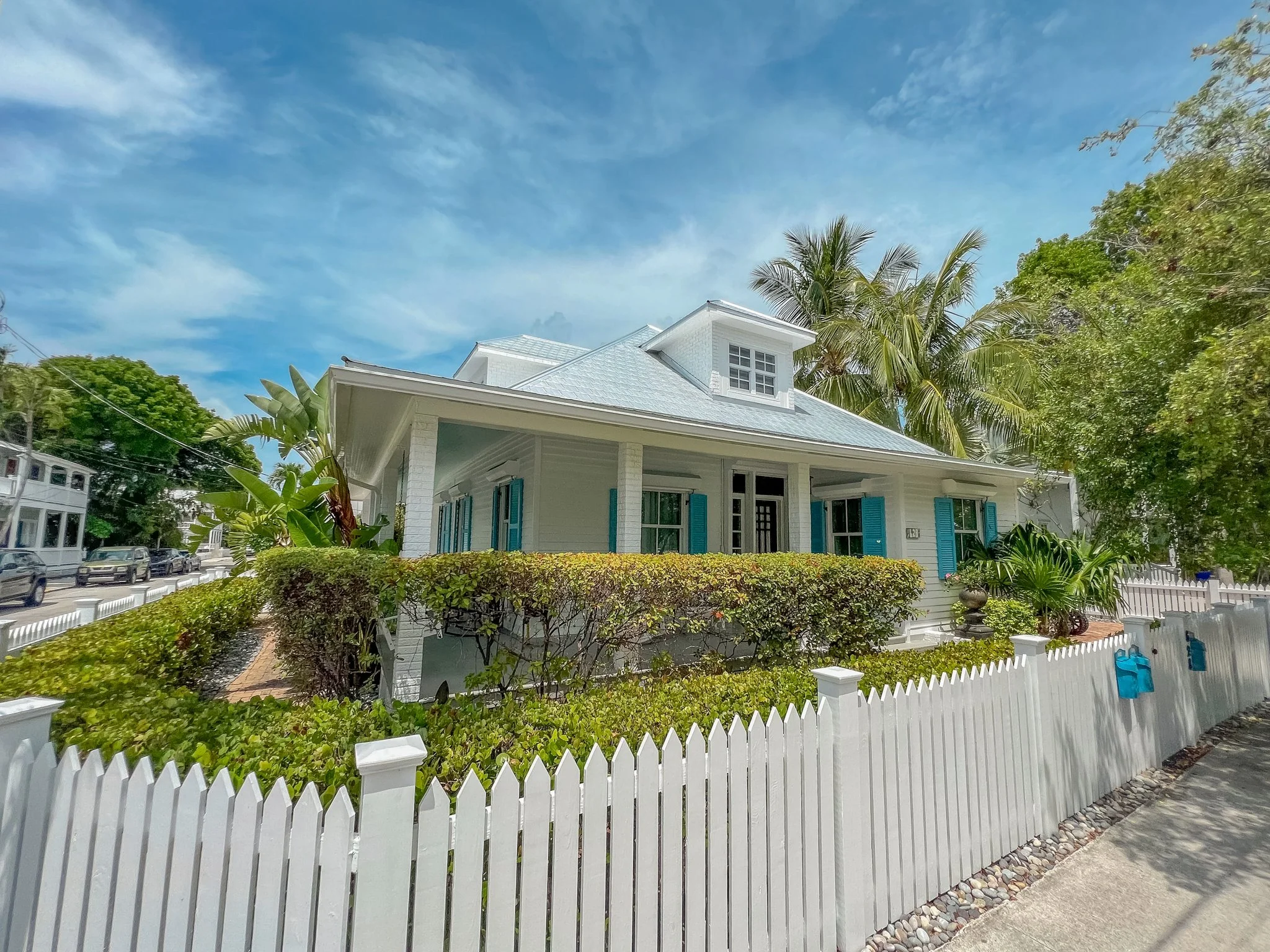 A white house with turquoise shutters and a metal roof surrounded by a white picket fence and lush greenery, with a sidewalk and cars on the street in front and a blue sky overhead.