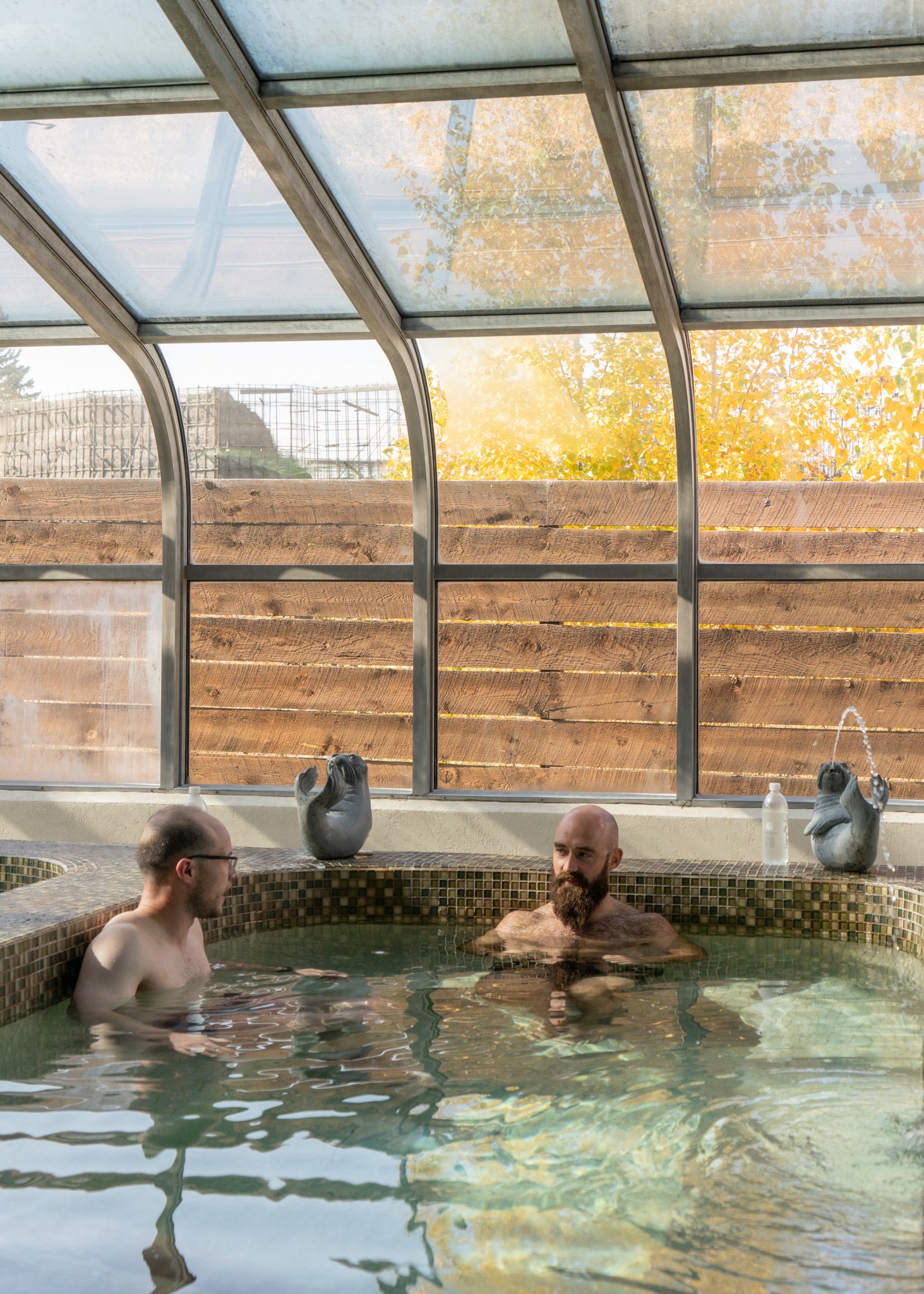 Two men relaxing in a tiled indoor hot tub with a glass ceiling and wooden wall outside, during autumn.