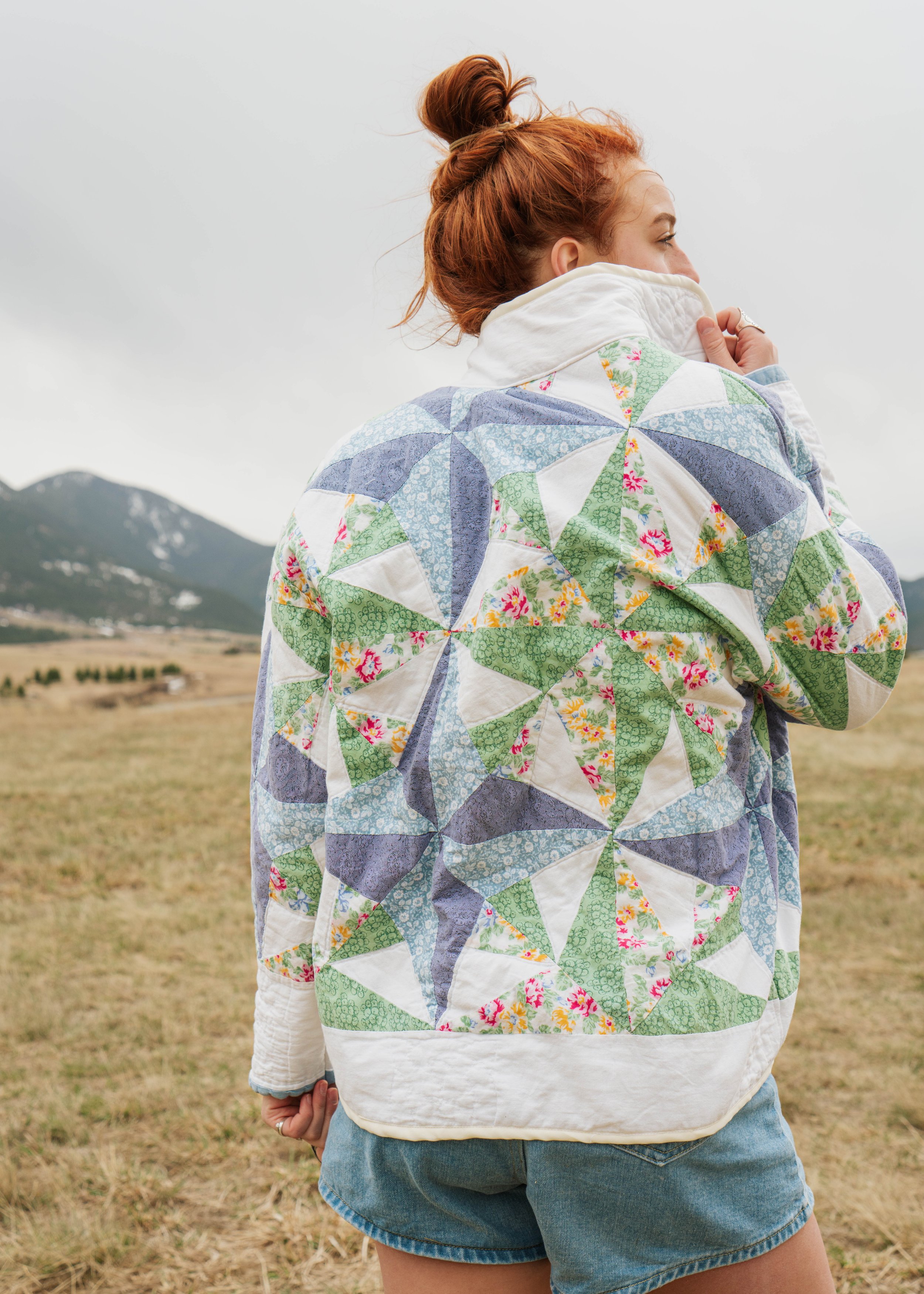 A woman with red hair in a bun walking outdoors in a field with mountains in the background, wearing a colorful quilted jacket with a patchwork pattern of green, blue, and floral fabrics.