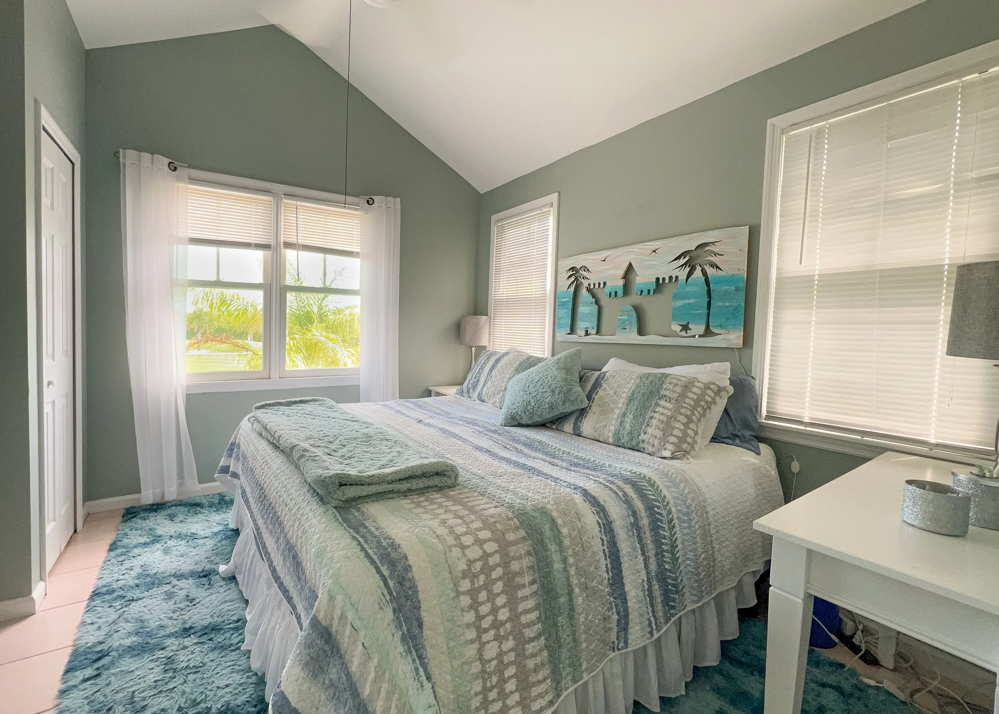 A cozy bedroom with light green walls, featuring a bed with patterned bedding and multiple pillows, a painting of a tropical scene above the headboard, two windows with blinds and sheer curtains, and a white nightstand with a lamp.