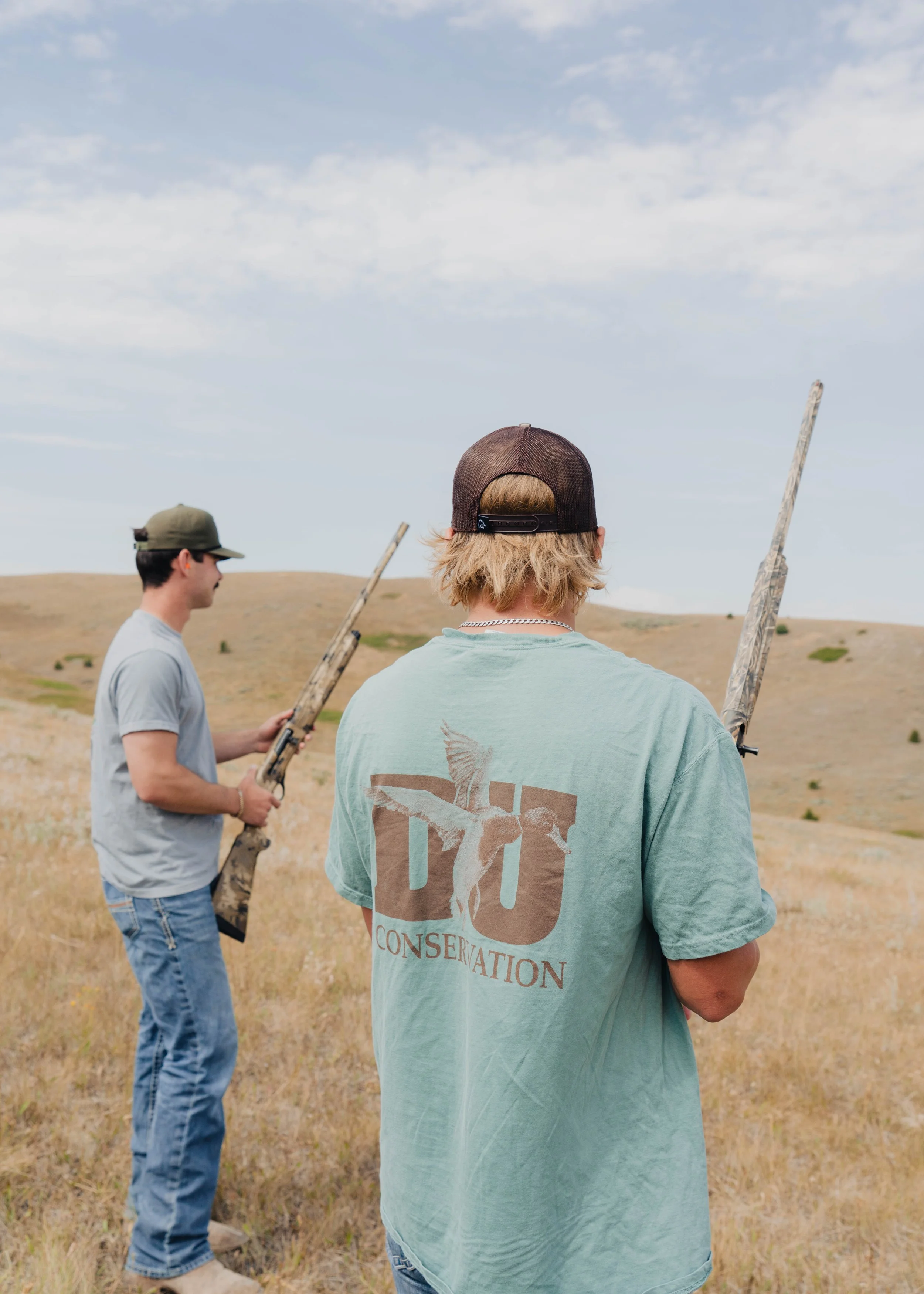 Two young men outdoors in a field, holding rifles, with rolling hills and partly cloudy sky in the background. One man is facing away, wearing a green T-shirt with a conservation logo. The other is in profile, wearing a gray T-shirt and a cap.