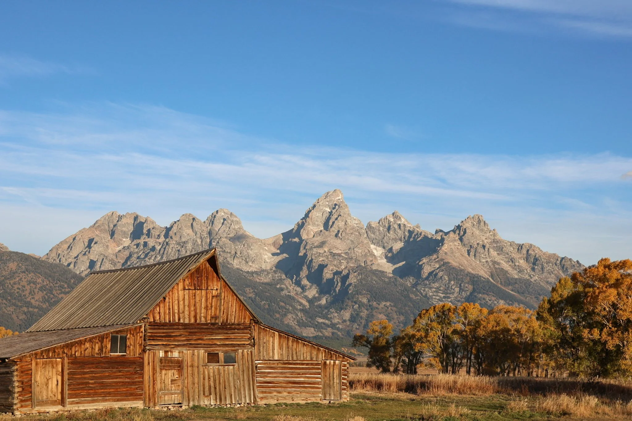 An old wooden barn with a slanted roof in a field with tall grass, trees with orange and green foliage, and mountains in the background under a blue sky with wispy clouds.