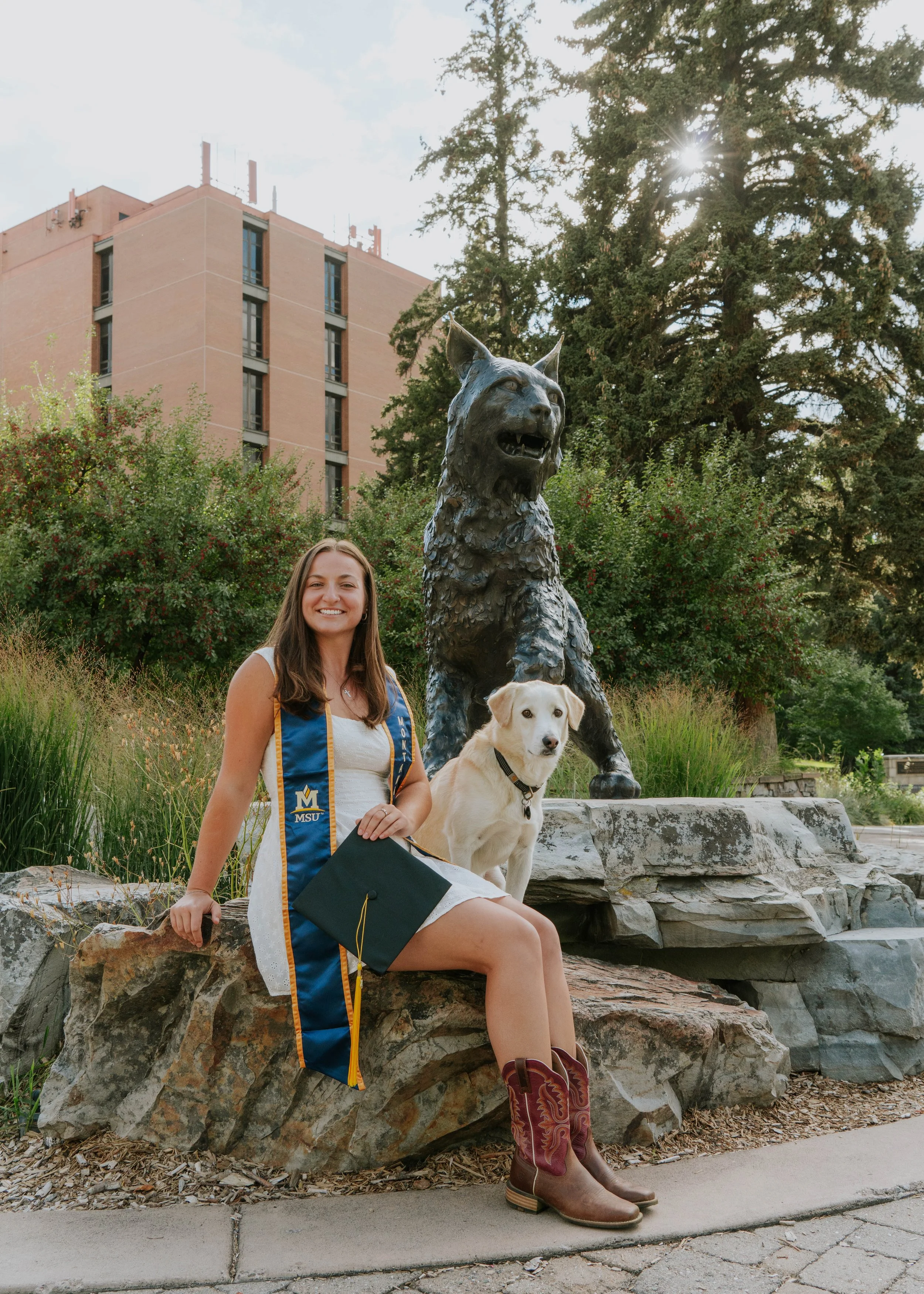 A young woman in a white dress and cowboy boots sitting on a rock, wearing a blue graduation stole, holding a cap, with a dog beside her, in front of a statue of a wolf on a college campus with trees and a building in the background.