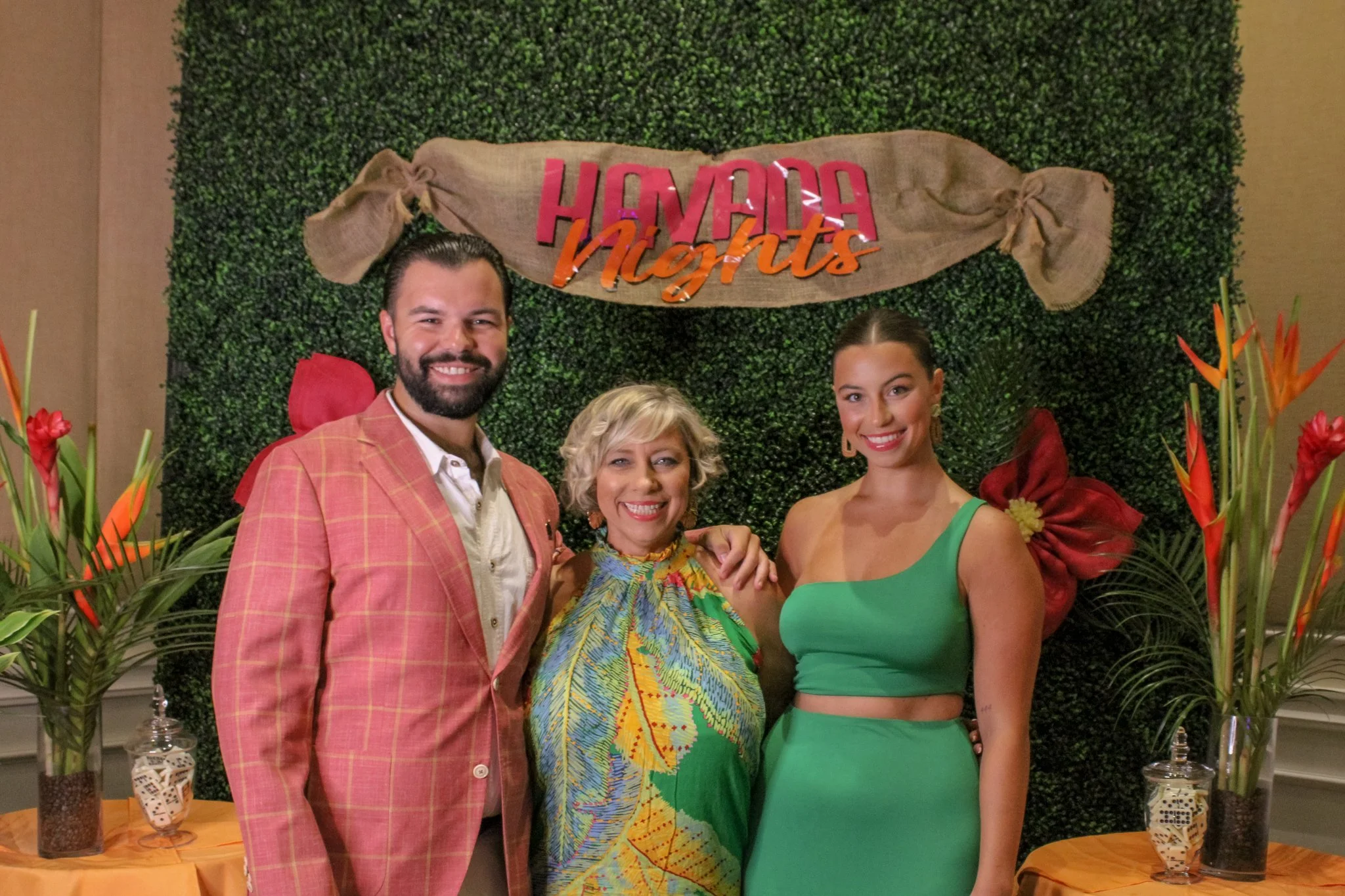 A group of three people smiling and posing in front of a decorative green hedge wall with a banner that reads 'Havana Nights'. There are floral arrangements on tables on each side of them.