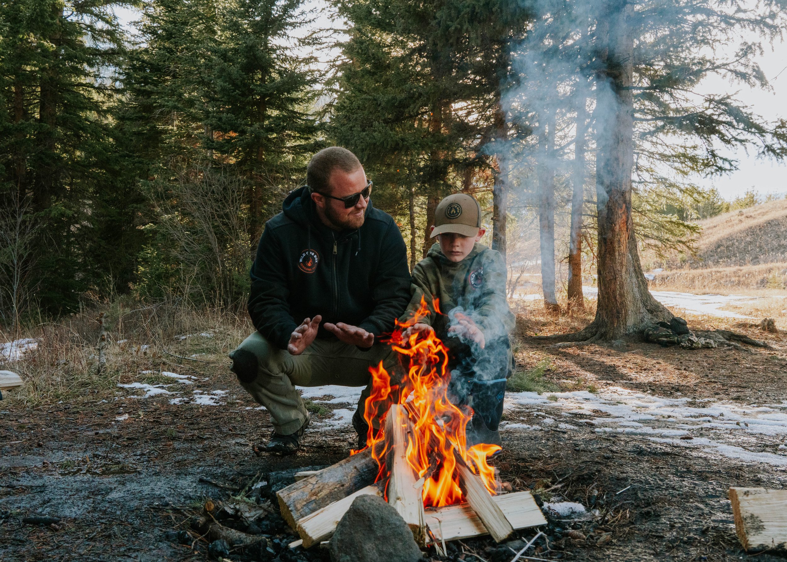 A man and young boy sitting by a campfire in a forest, with pine trees and some patches of snow on the ground.