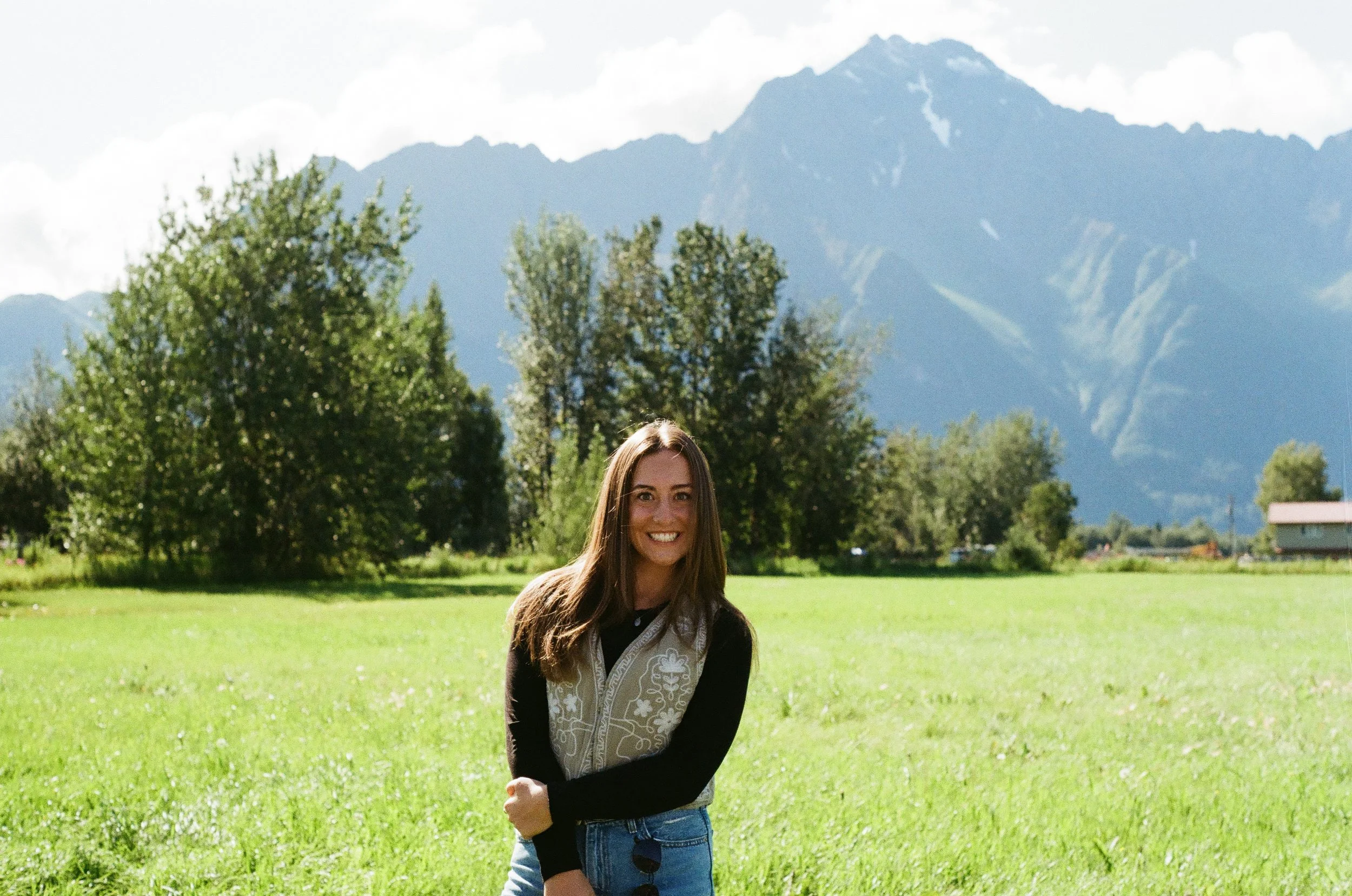 A smiling woman standing in a grassy field with trees and mountains in the background.